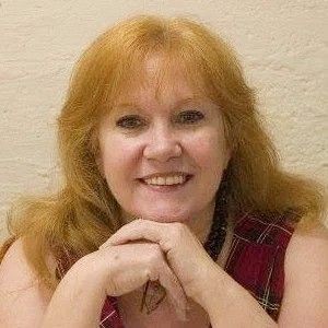 Woman with reddish-blonde hair, hands clasped, smiling, wearing a plaid top.