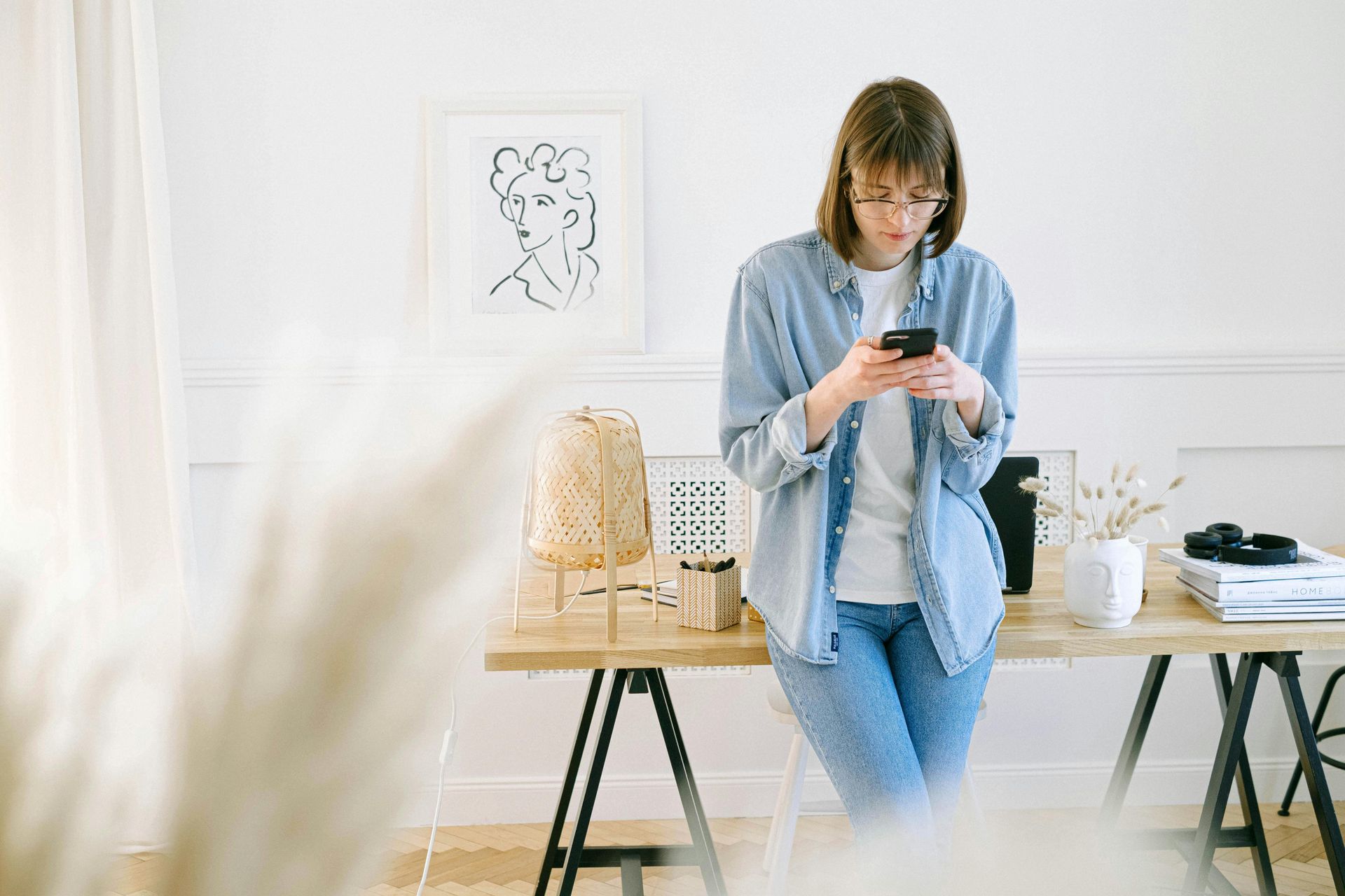 Woman in jeans and a denim shirt looks at her phone while leaning on a desk in a bright room.