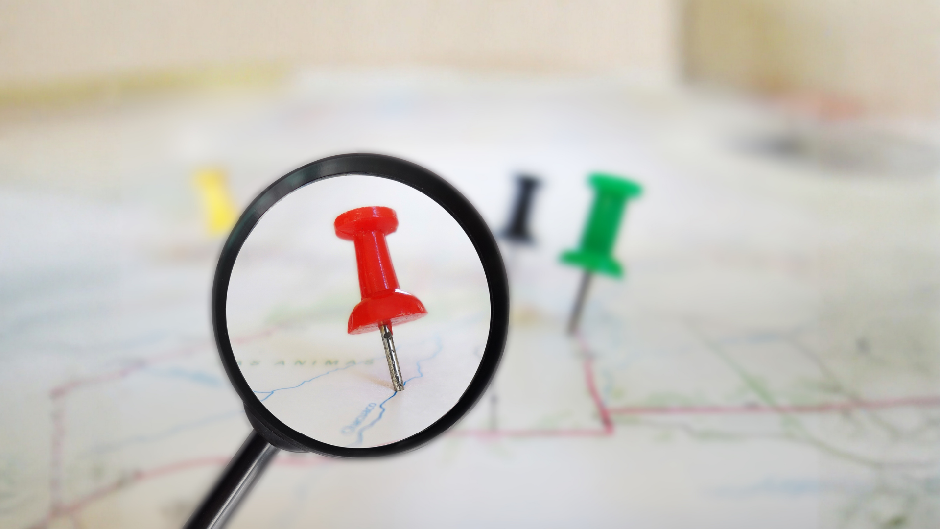 Magnifying glass over a red pushpin on a map with a green pushpin in the background