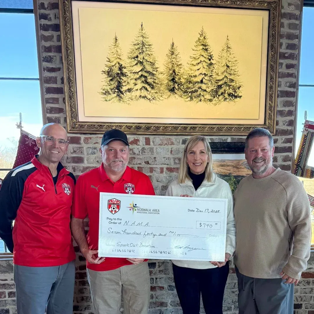 Four people stand in front of a brick wall holding a donation check in a room with a painting of pine trees.