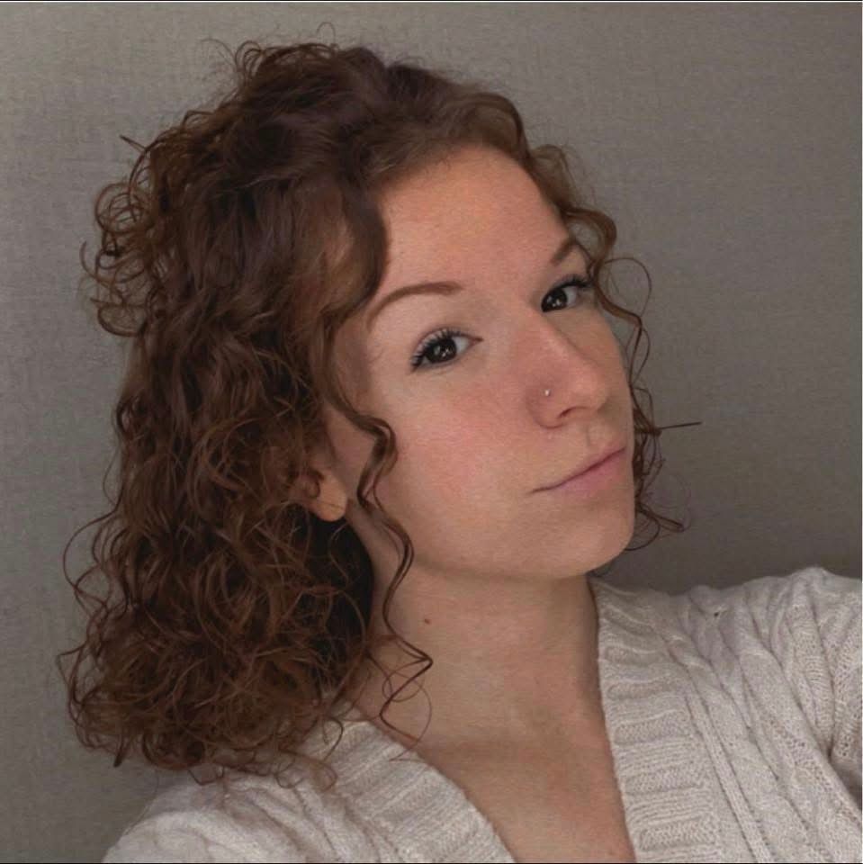 A woman with curly hair is wearing a white sweater and taking a selfie.