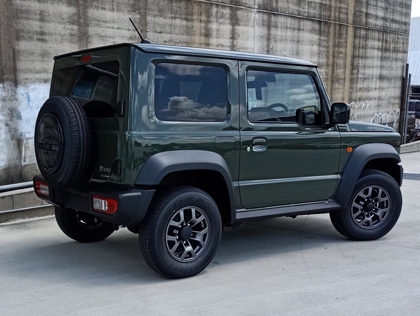 A green suzuki jimny is parked in front of a concrete wall.