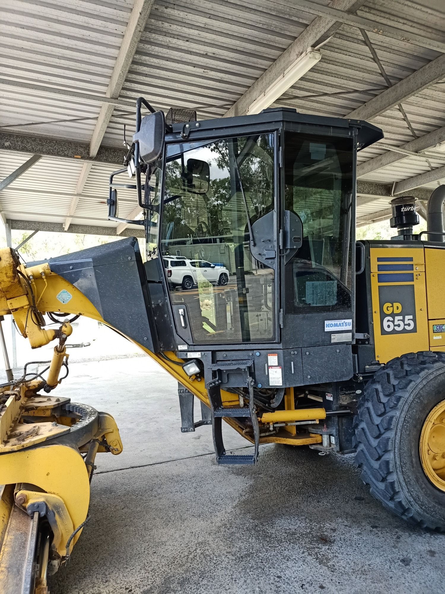Motor grader with a cab, parked under a shelter. Yellow and black machine.
