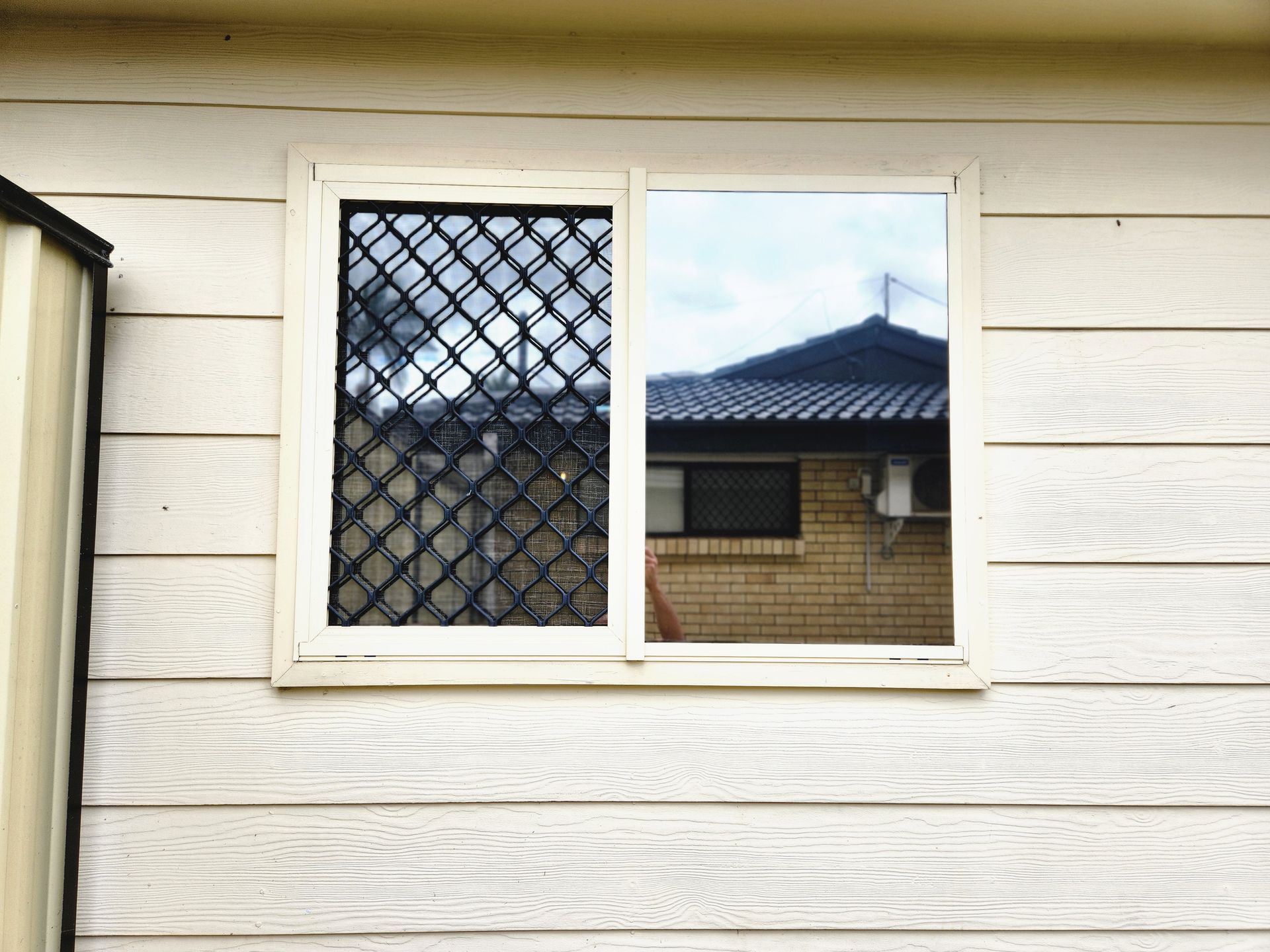 Window with security screen on the left, reflecting a house with a dark roof and brick wall on the right.