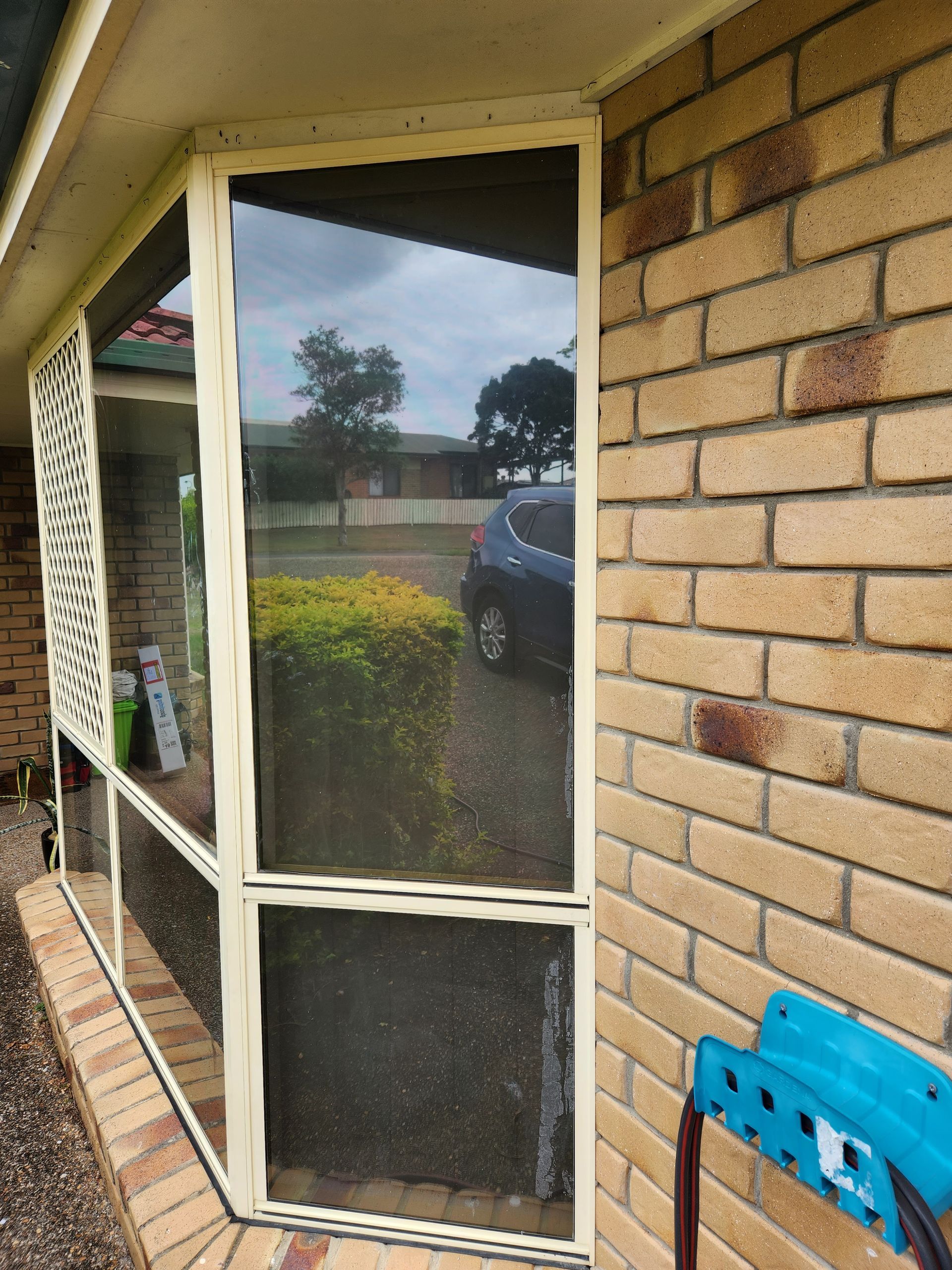 Window with cream frame and tinted glass next to a brick wall. A car and bushes are visible through the glass.