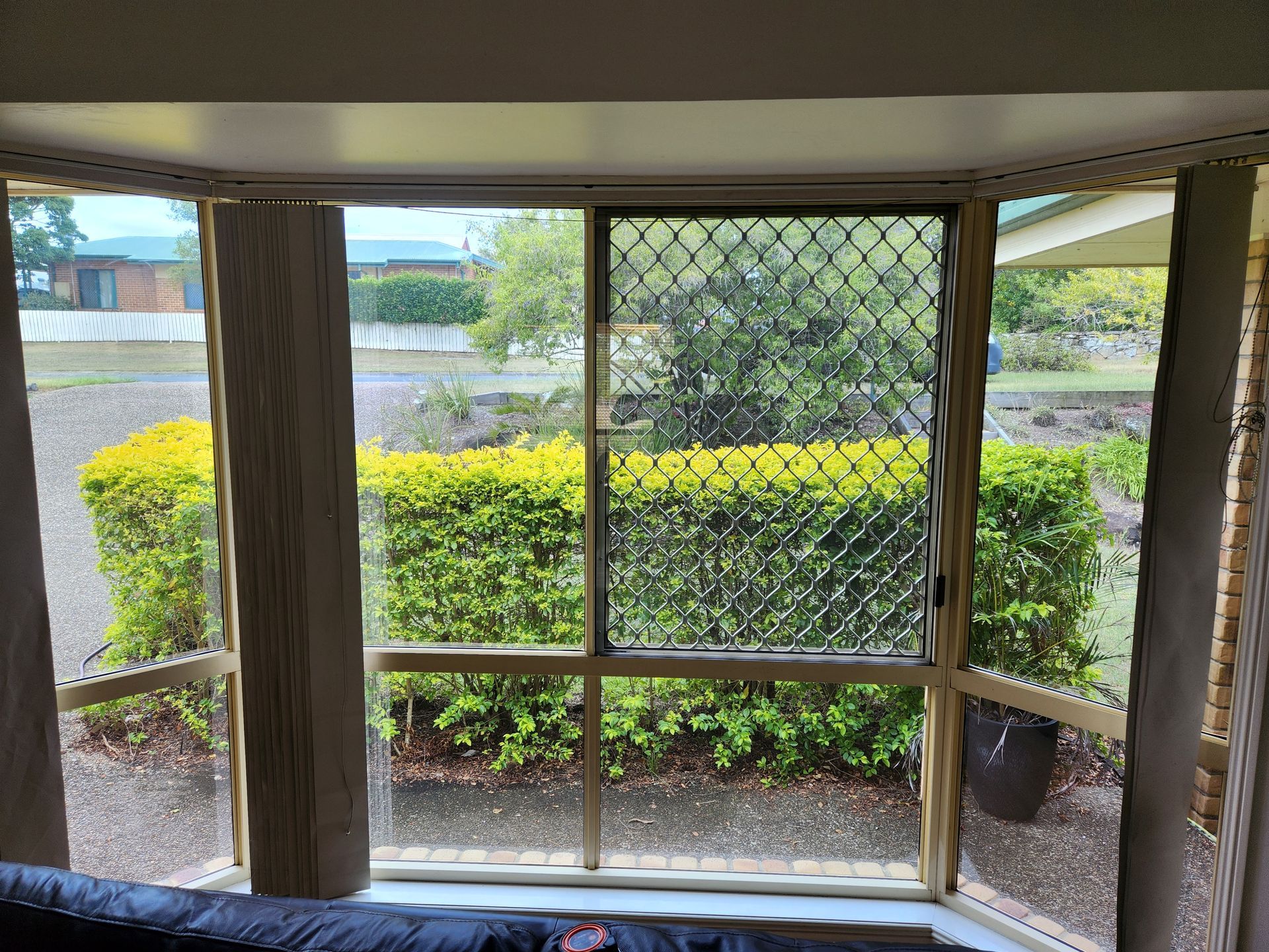 Bay window overlooking a green hedge.  Closed security screen and beige curtains frame the view.