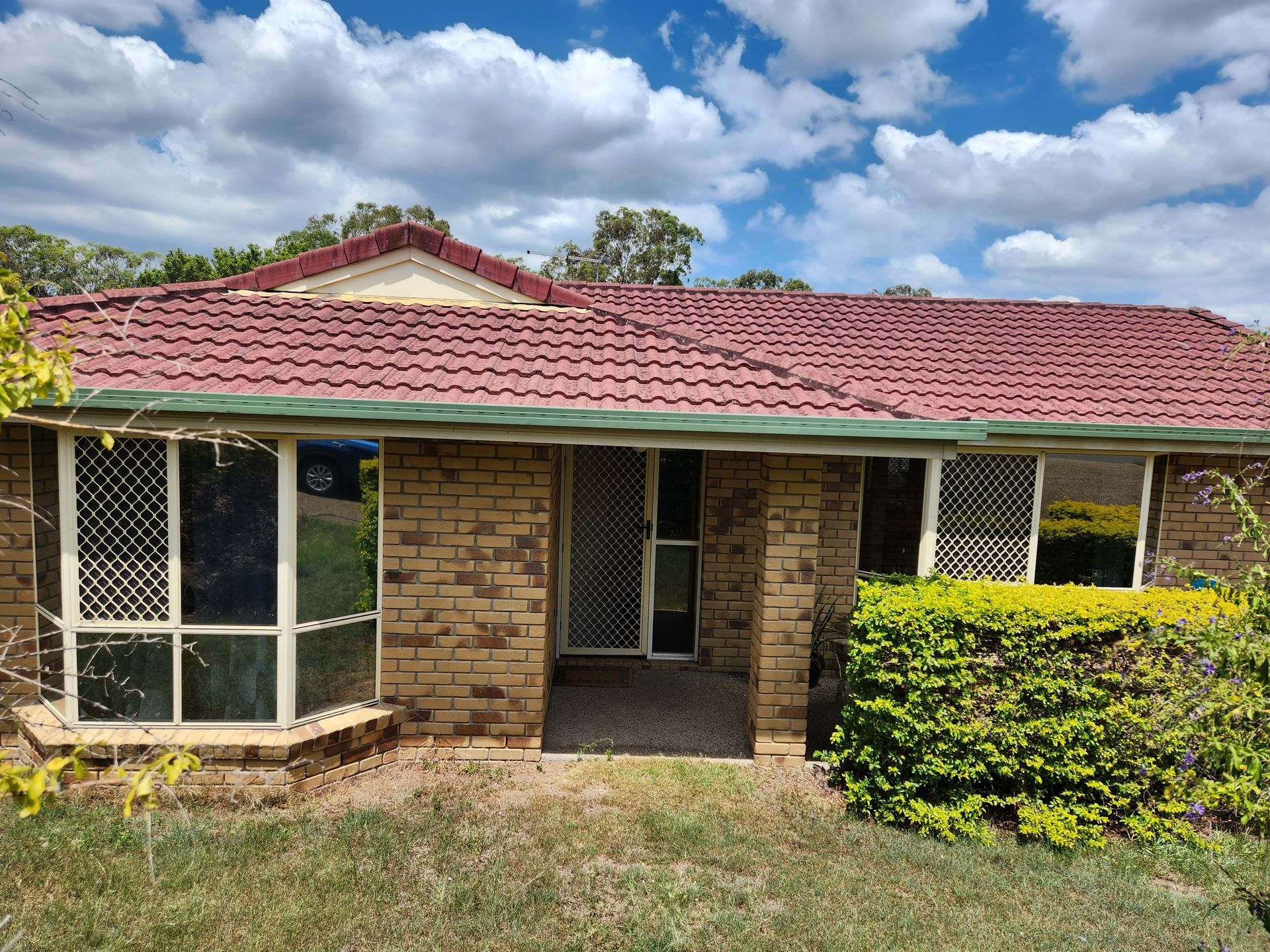 Brick house with red tile roof, security screens on windows. Cloudy blue sky overhead.
