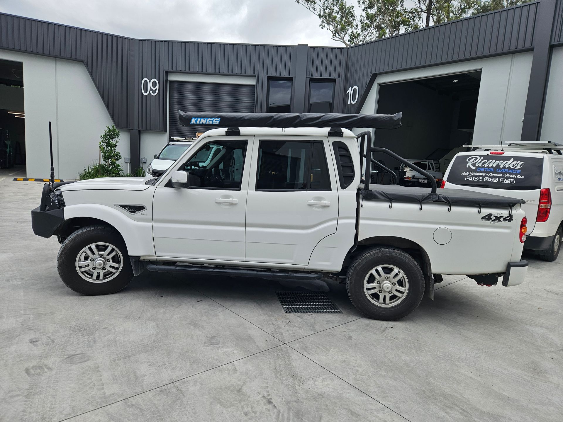 White pickup truck parked outside a building with an open garage door and a commercial van nearby.
