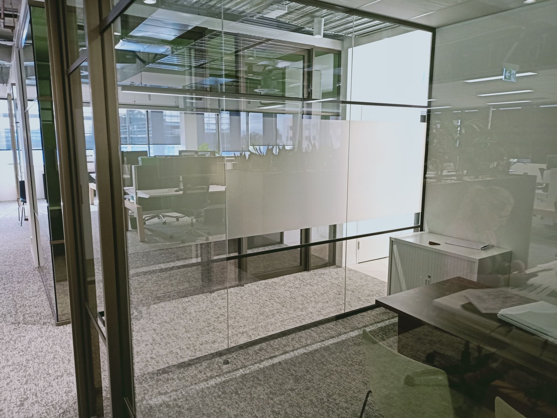 Modern office with glass walls, frosted privacy panels, and a desk. Sunlight filters in.