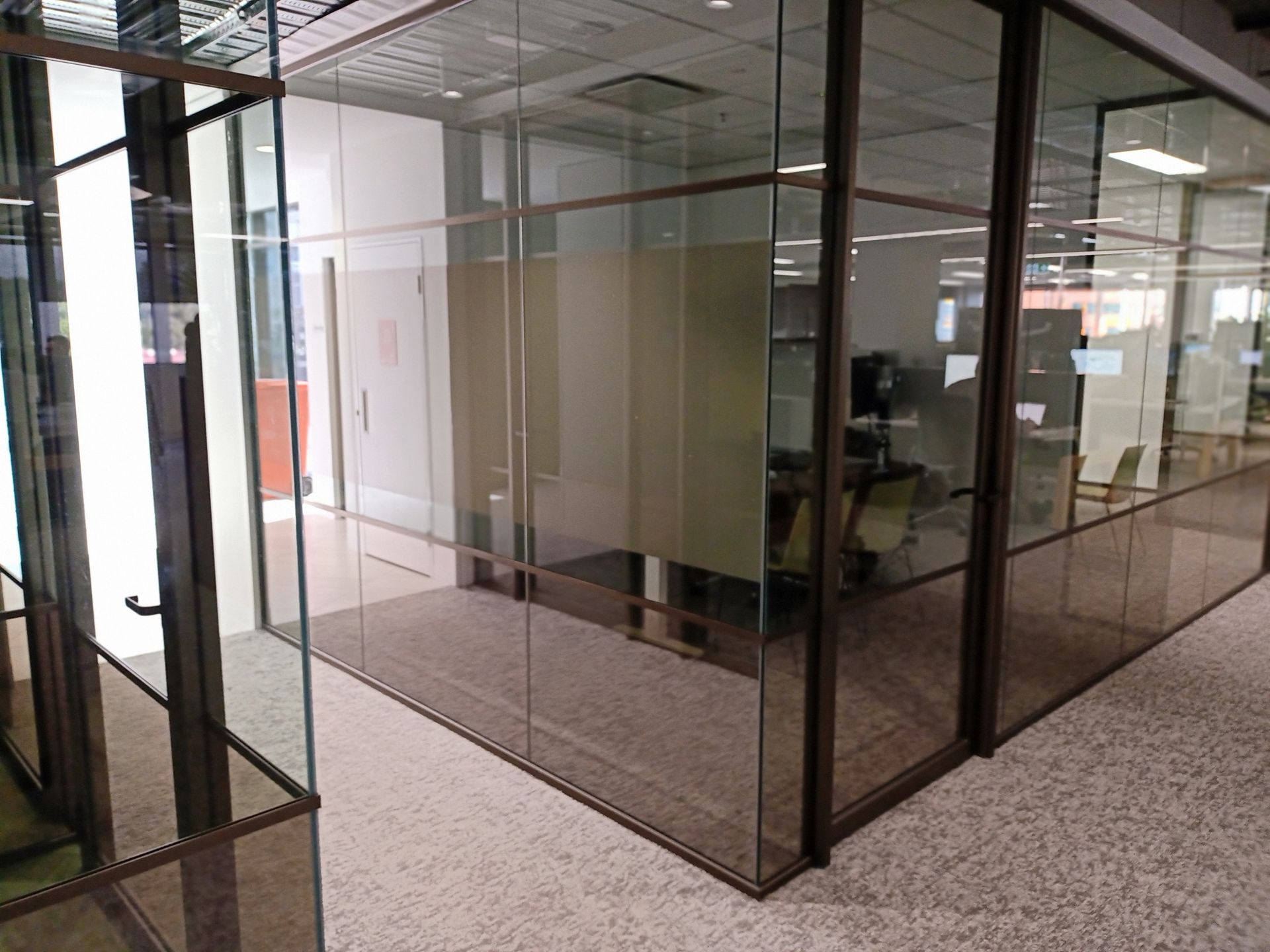 Glass-walled office cubicle with tinted windows and brown frames, in a brightly lit office space.