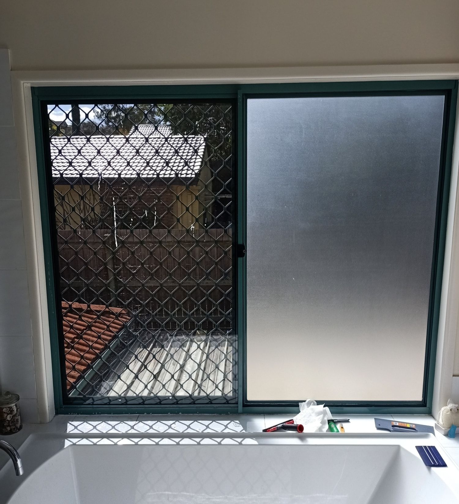 Bathroom window with security screen on the left, frosted glass on the right, overlooking a rooftop and backyard.