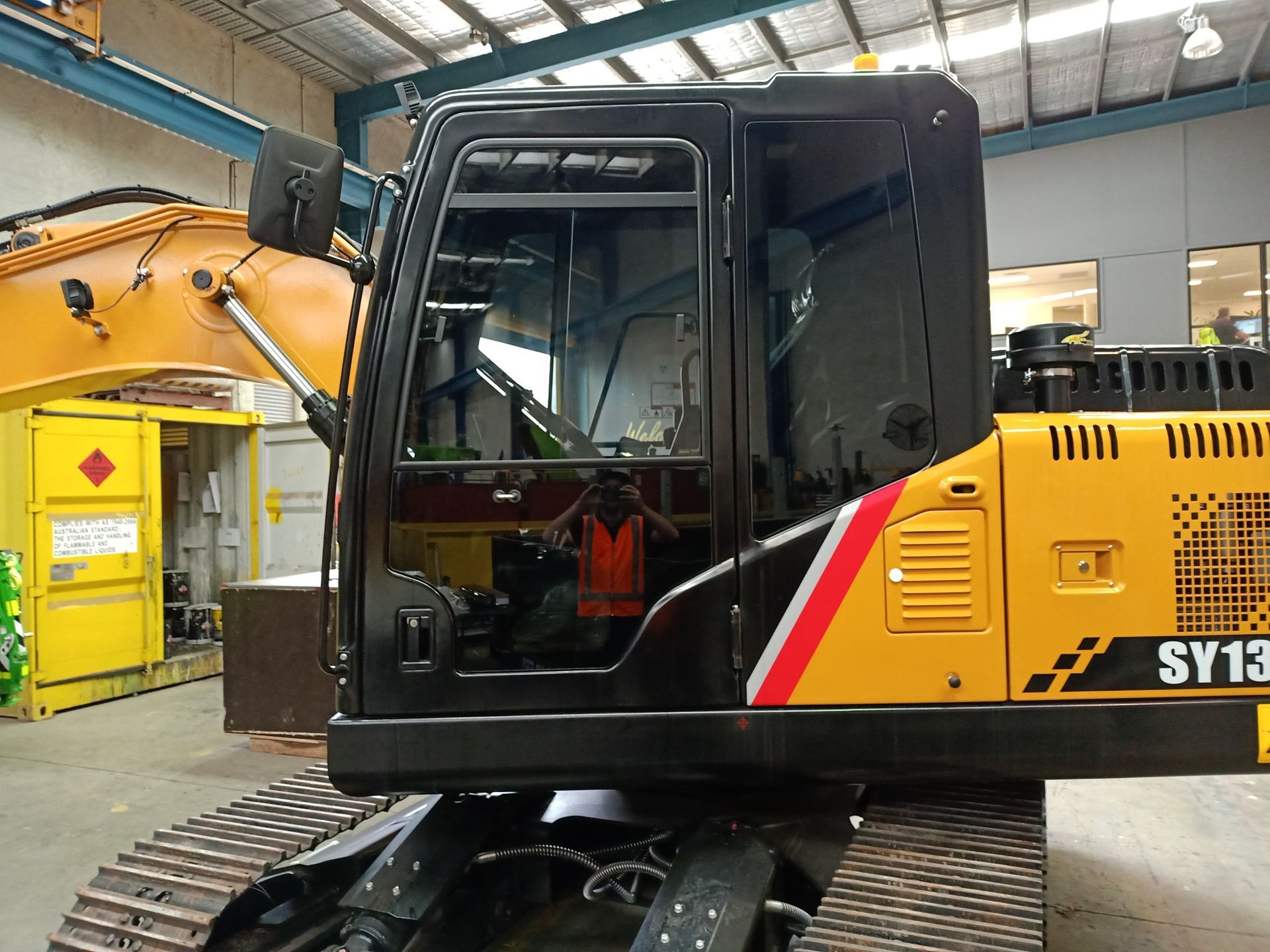 Black and yellow excavator cab with person in orange vest reflected in window.