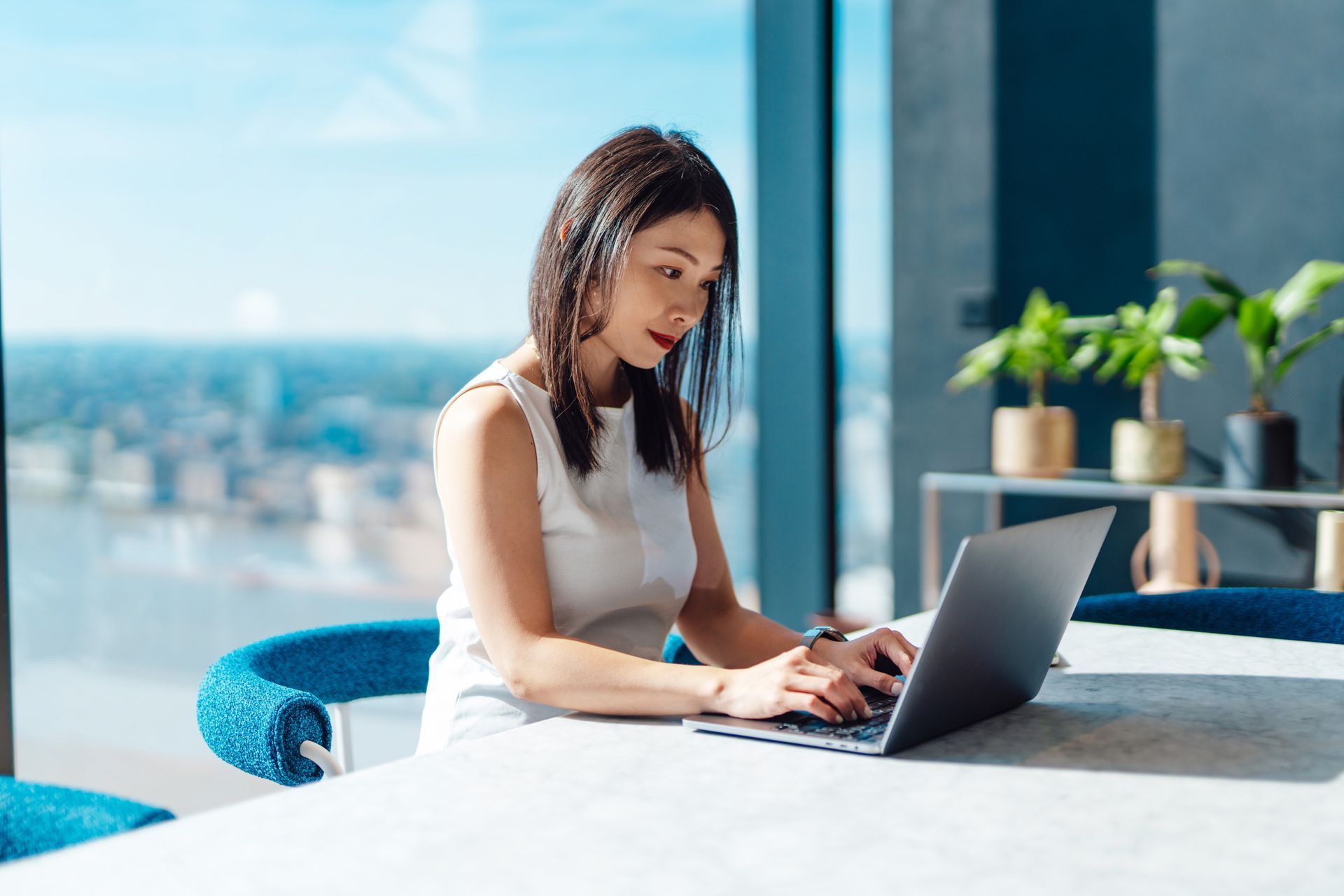 Asian woman working with a laptop while sitting by the window with city view and sunlight.