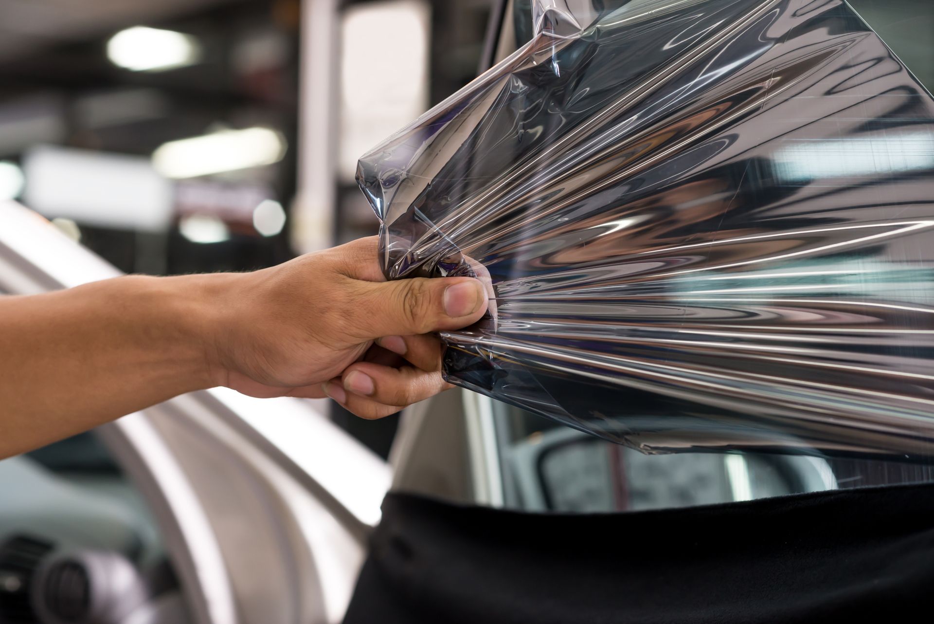 A man’s hand removes an old window tint from a car’s window by pulling on it.
