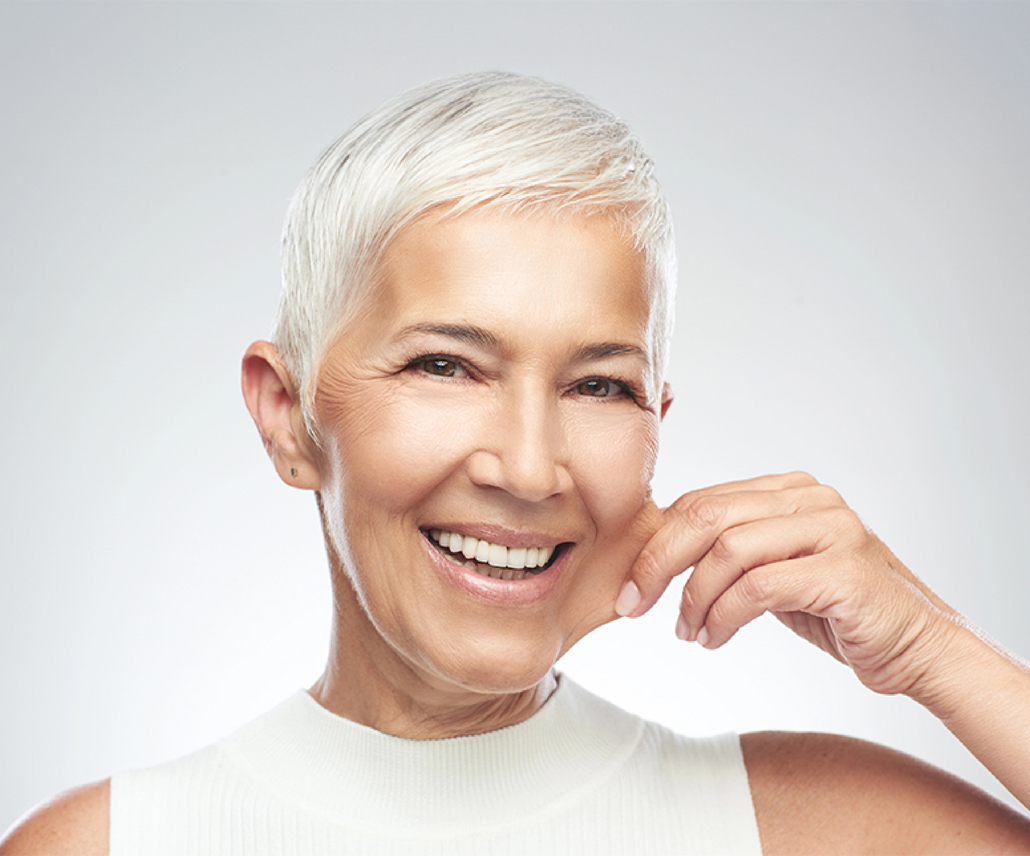 Eine Frau mit kurzen weißen Haaren lächelt und berührt ihr Gesicht.