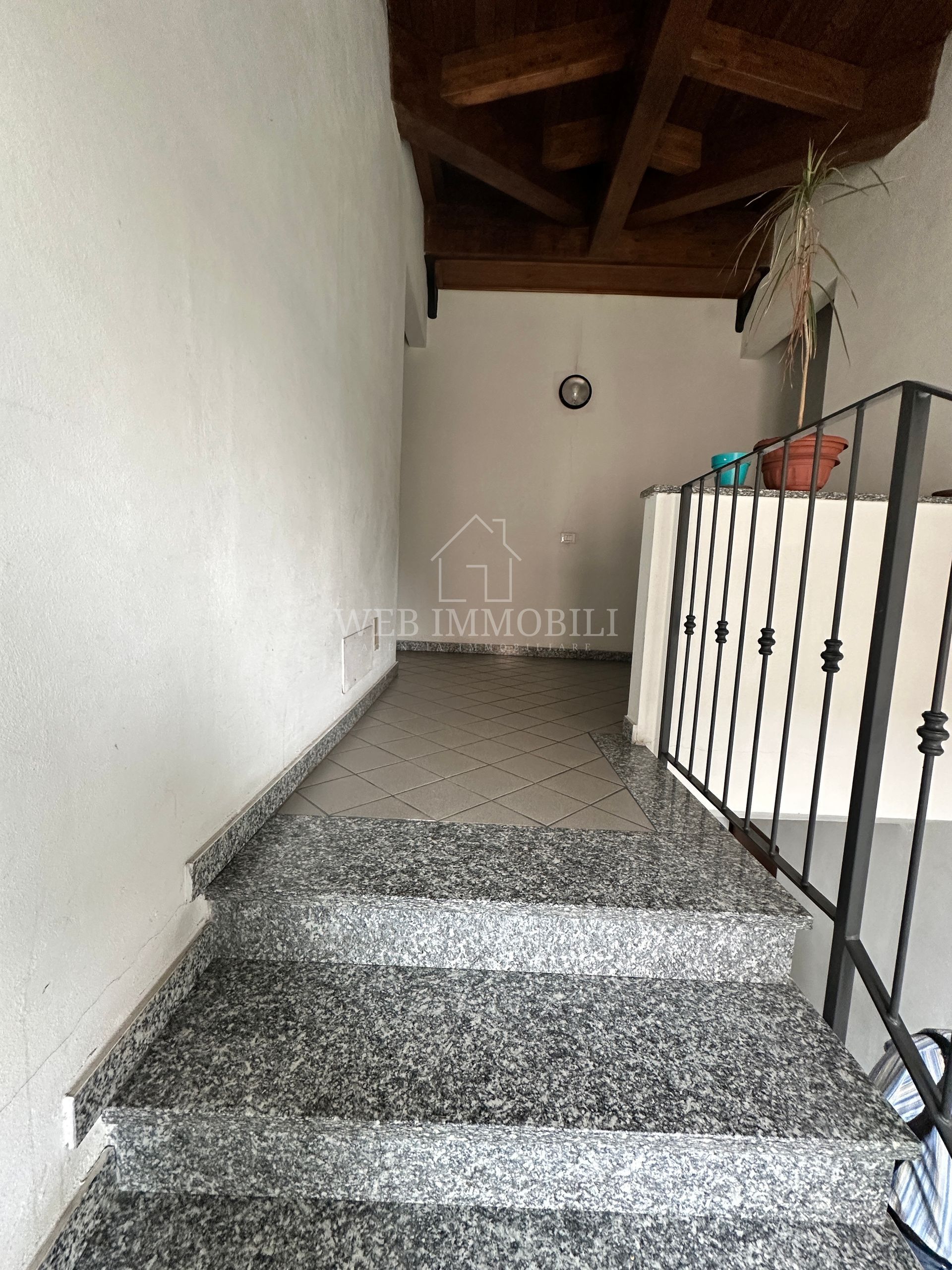 Scala in granito che conduce a un corridoio con pareti bianche, travi in ​​legno a vista e ringhiera in metallo.