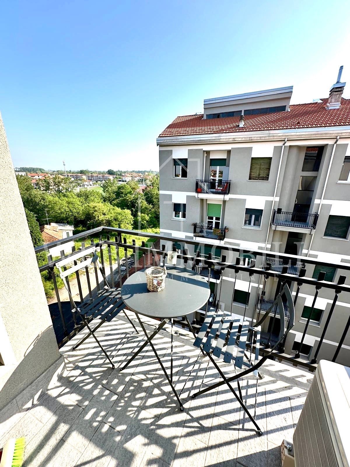 Balcone con tavolino e sedie, con vista sulla città, con alberi verdi sotto un cielo azzurro.
