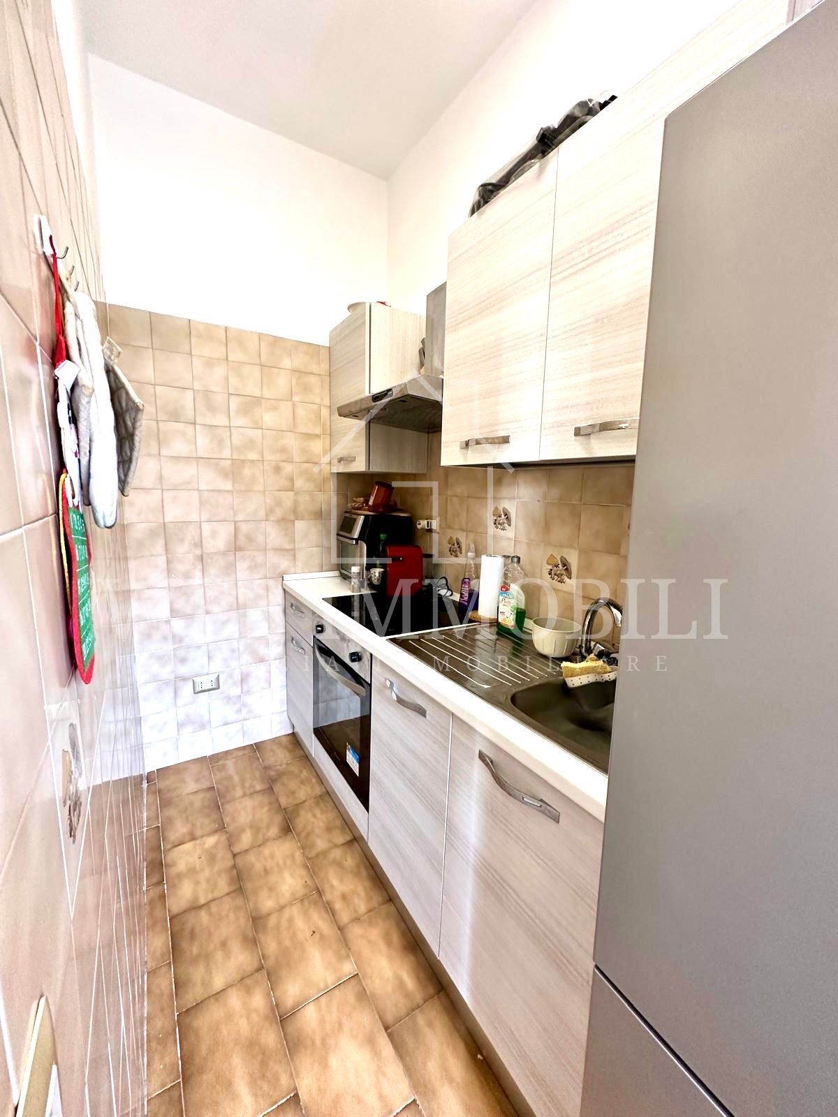 Cucina stretta con mobili in legno chiaro, stufa e paraschizzi piastrellato; prevalentemente toni neutri.