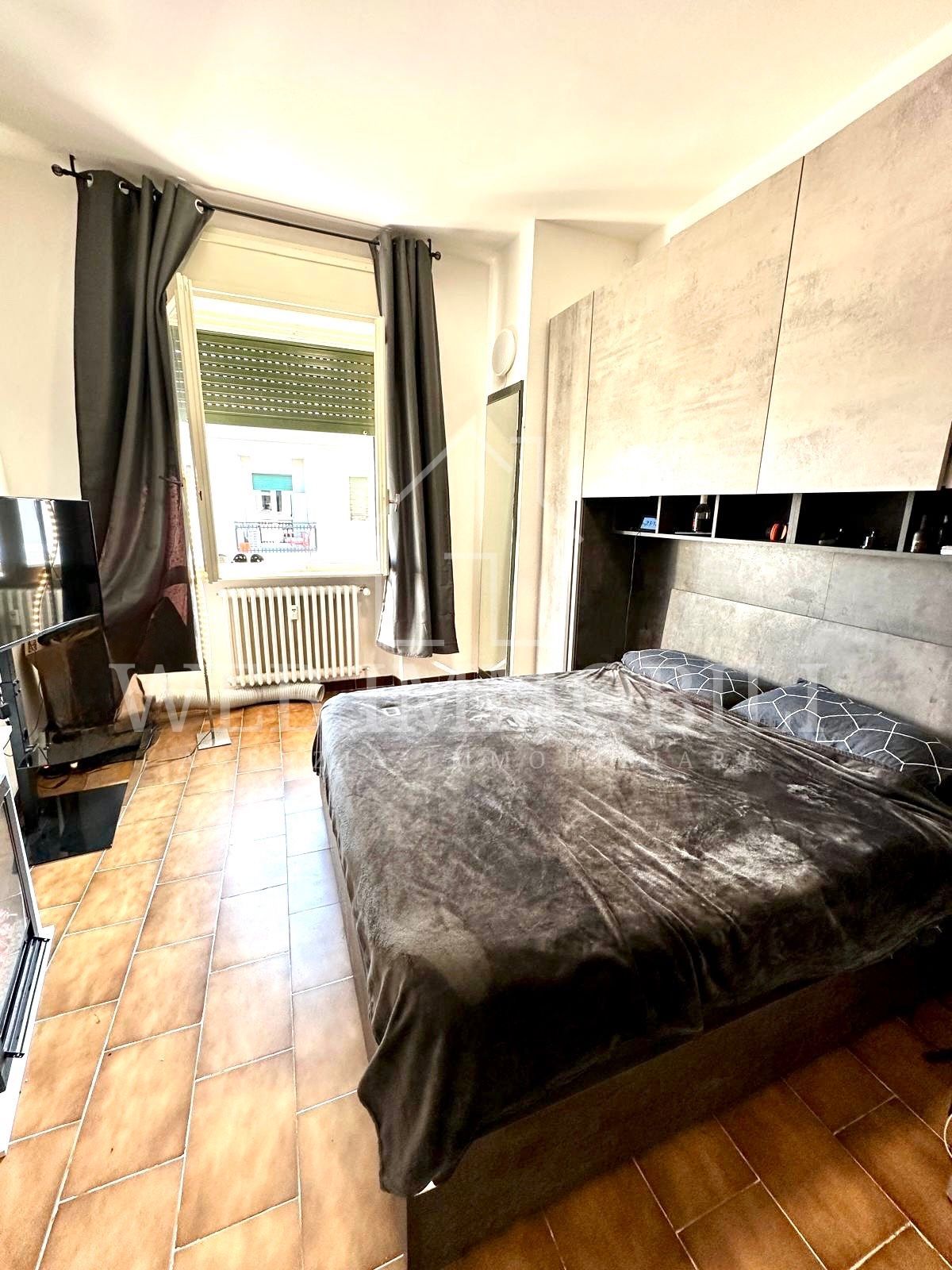 Camera da letto con letto, finestra con tende e scaffali a muro. Prevalentemente nei toni del grigio, con pavimento in piastrelle calde.