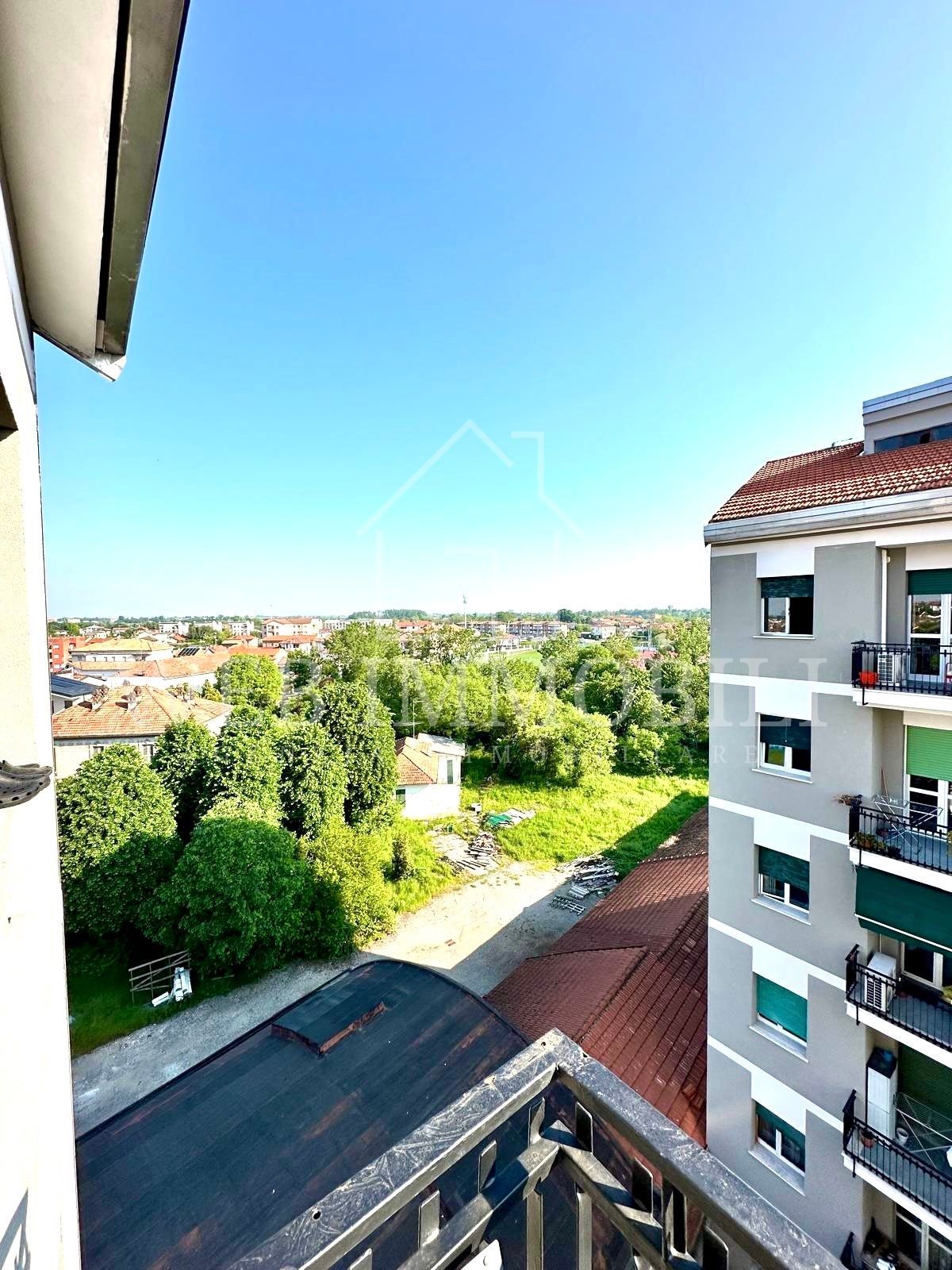 Vista da un balcone che domina una città con edifici, alberi e un cielo azzurro e limpido.