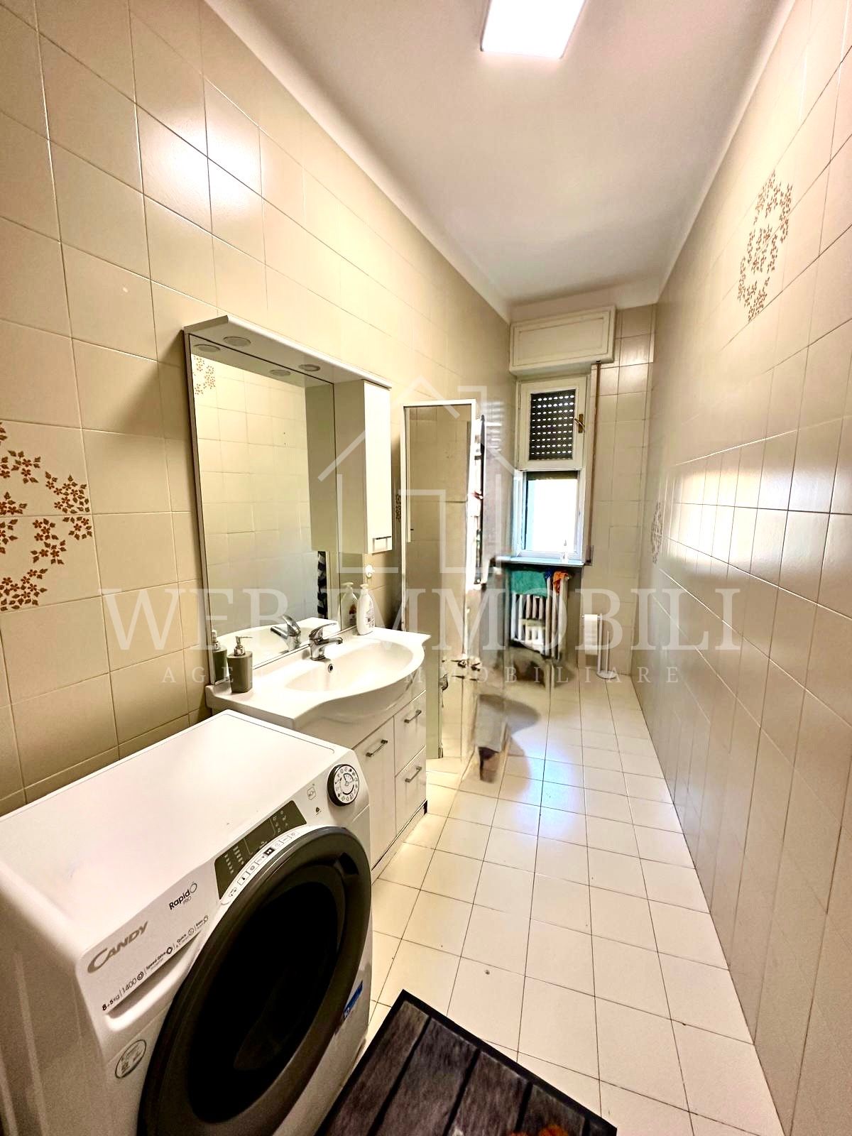 Piccolo bagno con lavatrice, lavandino, specchio, mobiletto e finestra, il tutto in toni neutri.