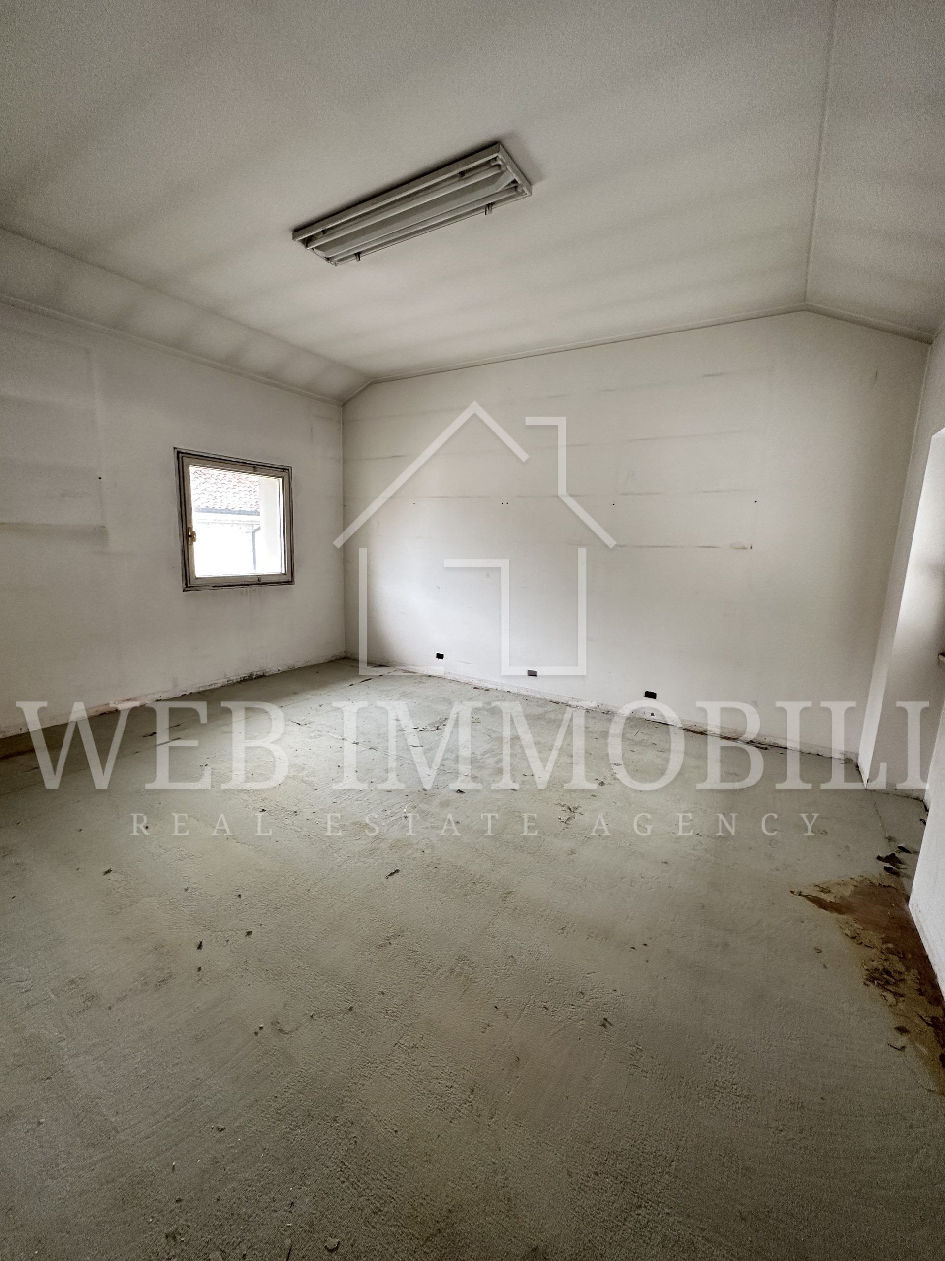 Stanza vuota, con pareti grigie, una piccola finestra e una lampada fluorescente sul soffitto.