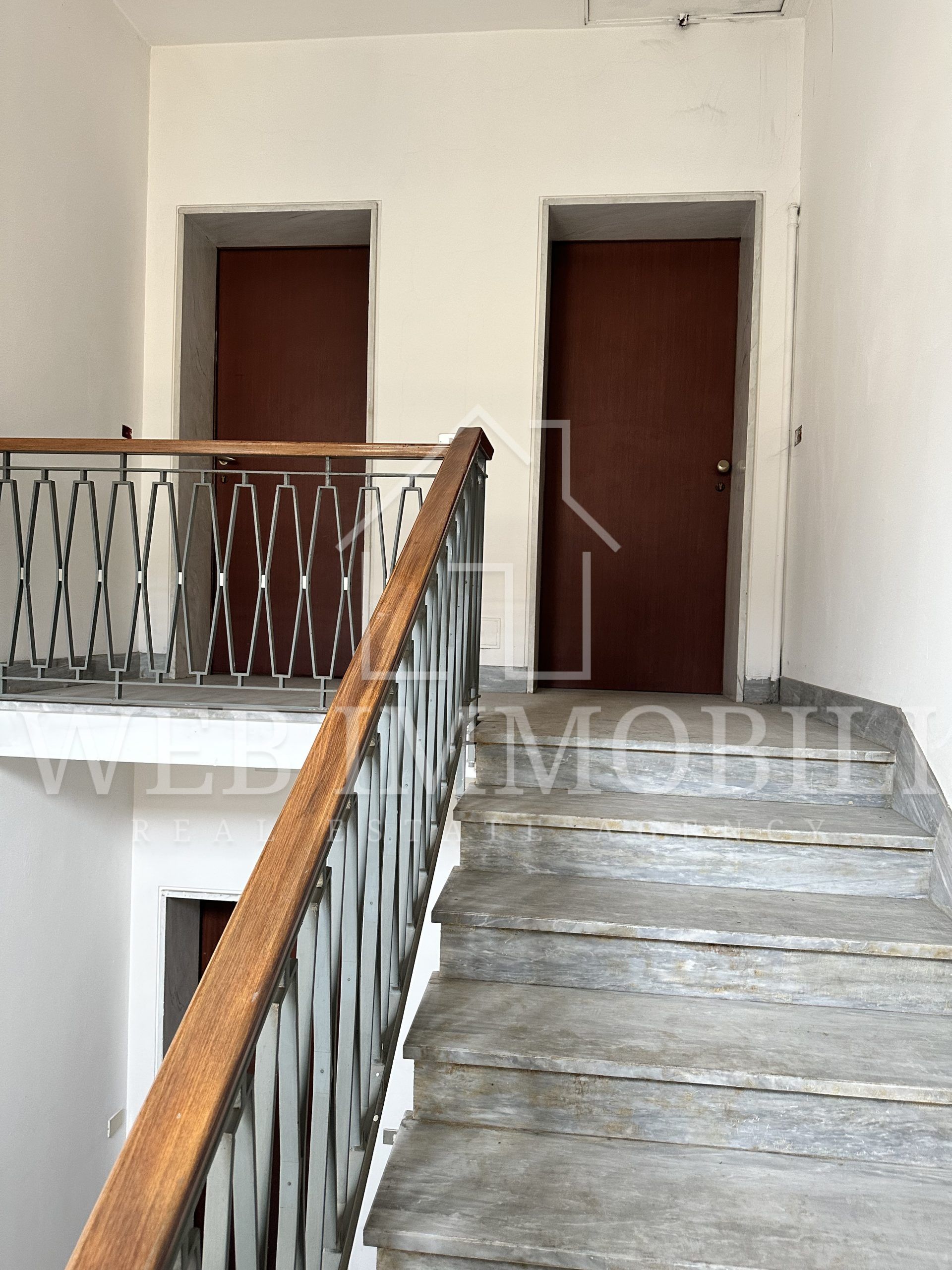 Tromba delle scale con gradini in cemento, porte marroni e ringhiera nera decorata con corrimano in legno.