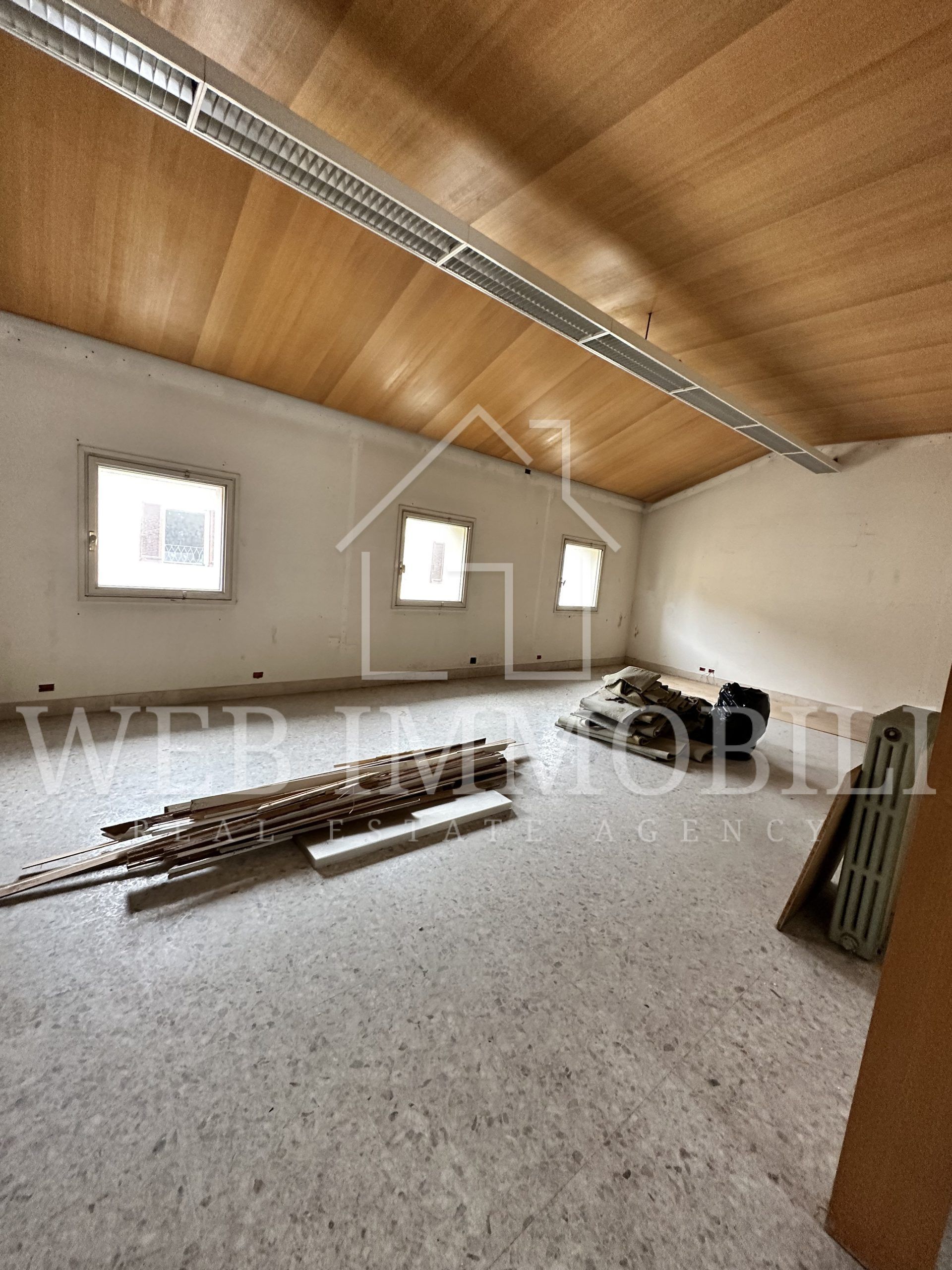 Vista interna di una stanza con pareti chiare, soffitto in legno, tre finestre e materiali da costruzione sul pavimento.