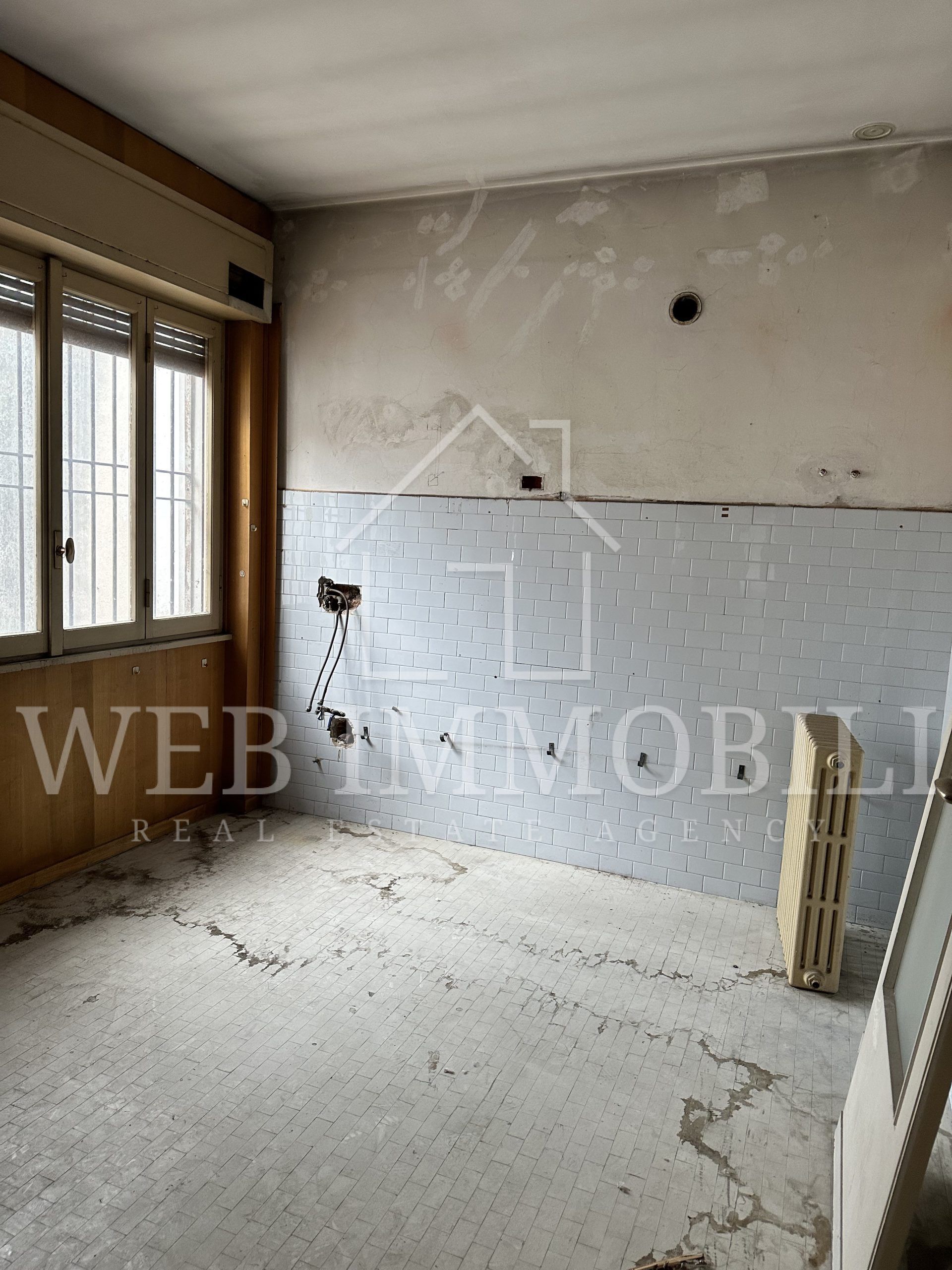 Una stanza vuota in fase di ristrutturazione: pavimento in cemento nudo, pareti parzialmente piastrellate, una finestra e detriti.