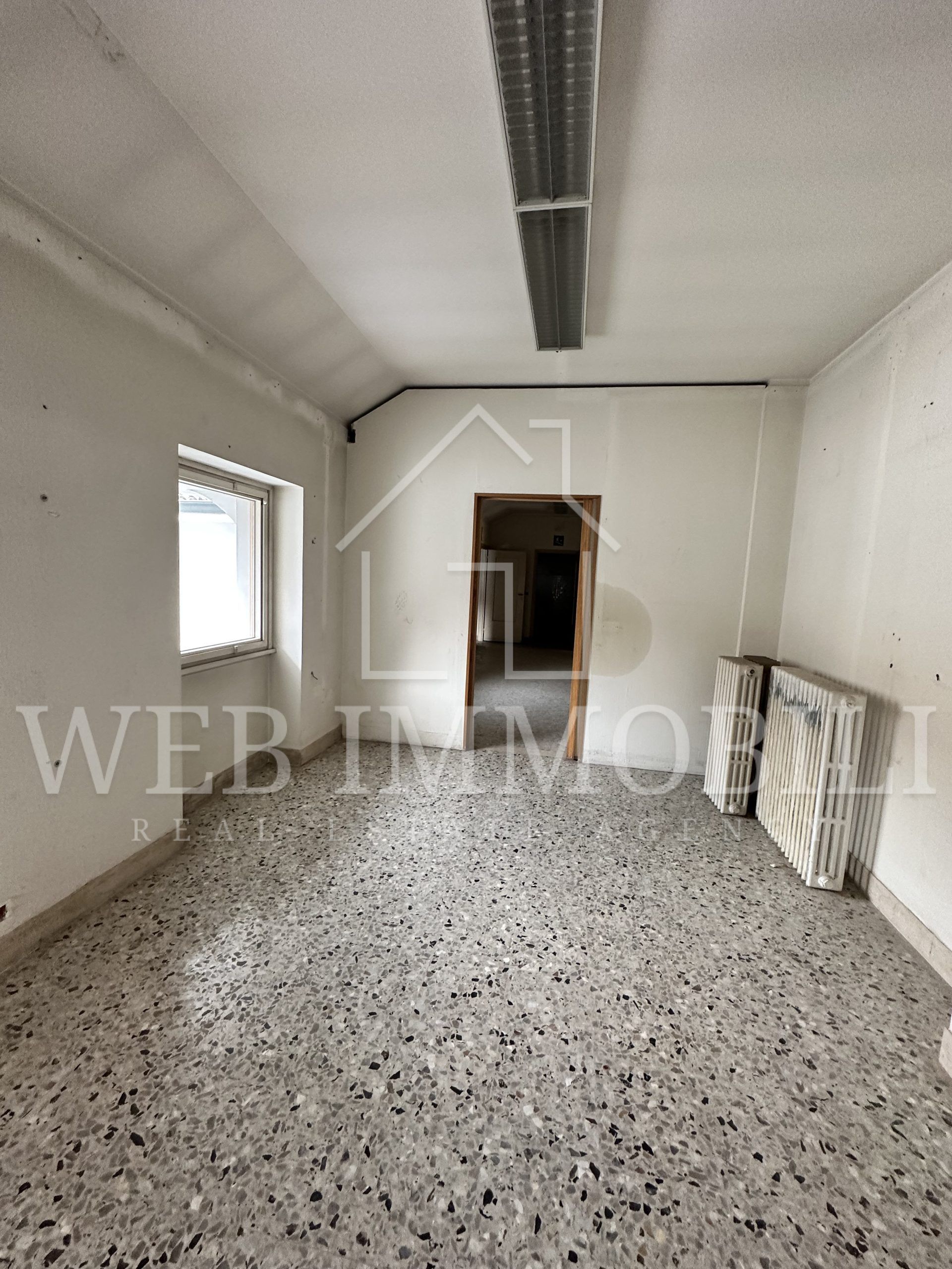 Stanza vuota con pavimento in terrazzo, piccola finestra, porta aperta e condotto del soffitto a vista.