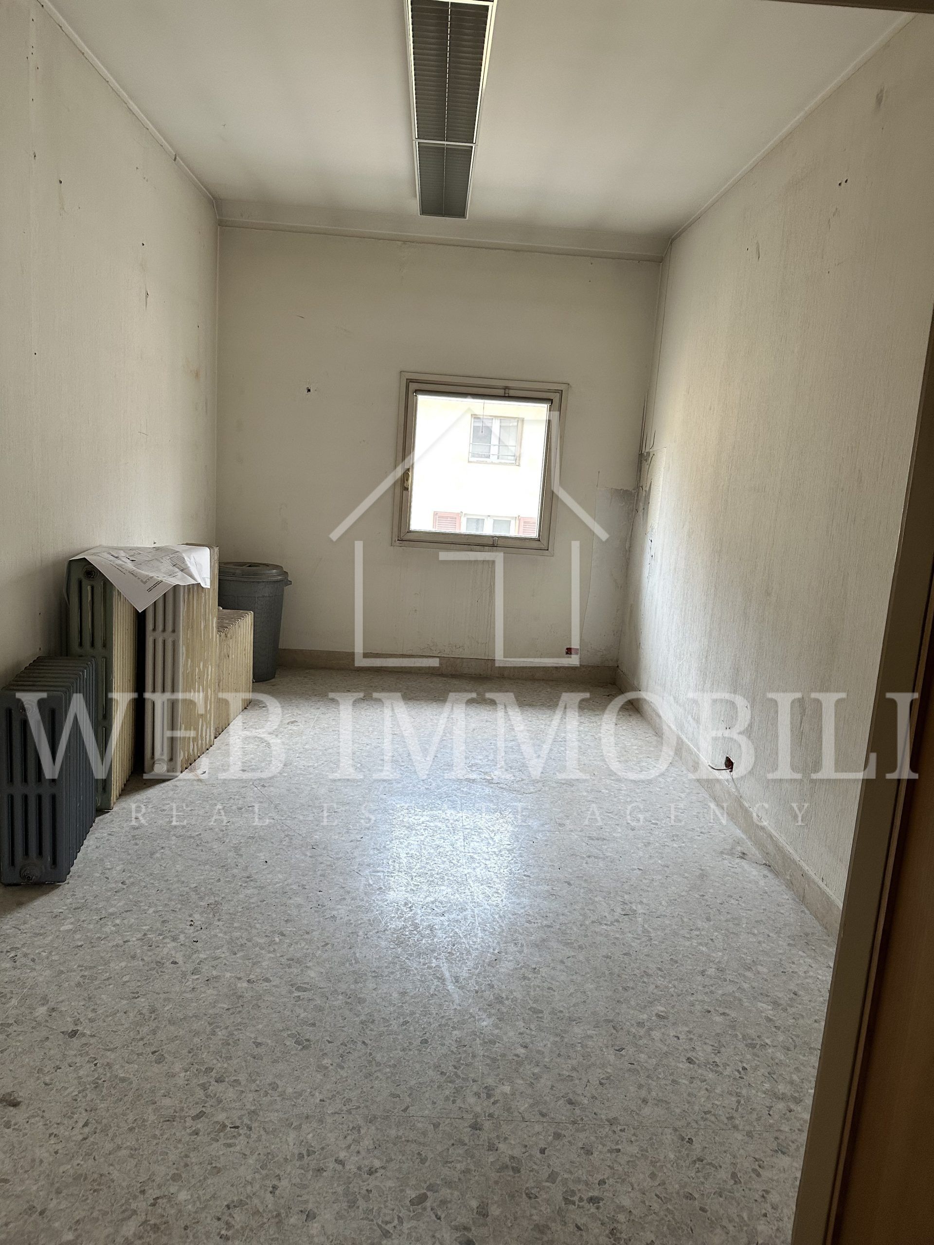 Stanza vuota con una finestra. Pavimento in terrazzo, pareti bianche, soffitto in cemento e materiali sulla sinistra.