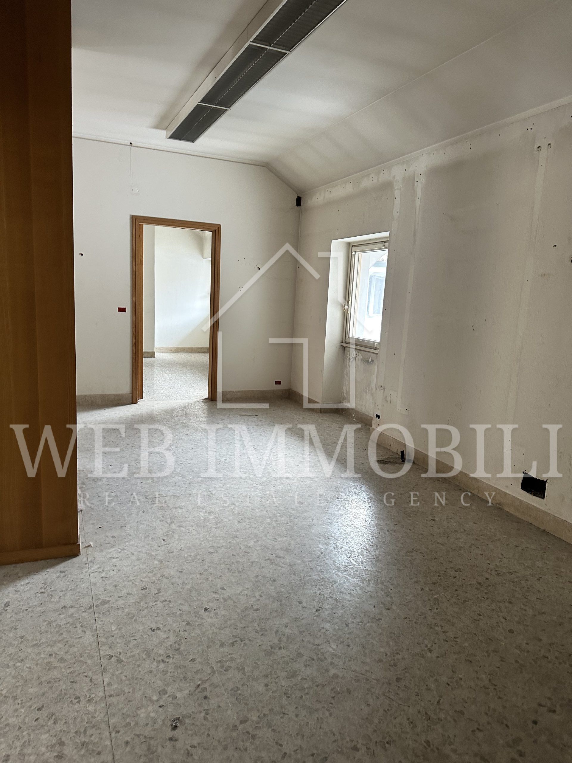 Stanza vuota con pareti chiare e pavimento in terrazzo, porta che conduce a un'altra stanza, finestra con tende bianche.