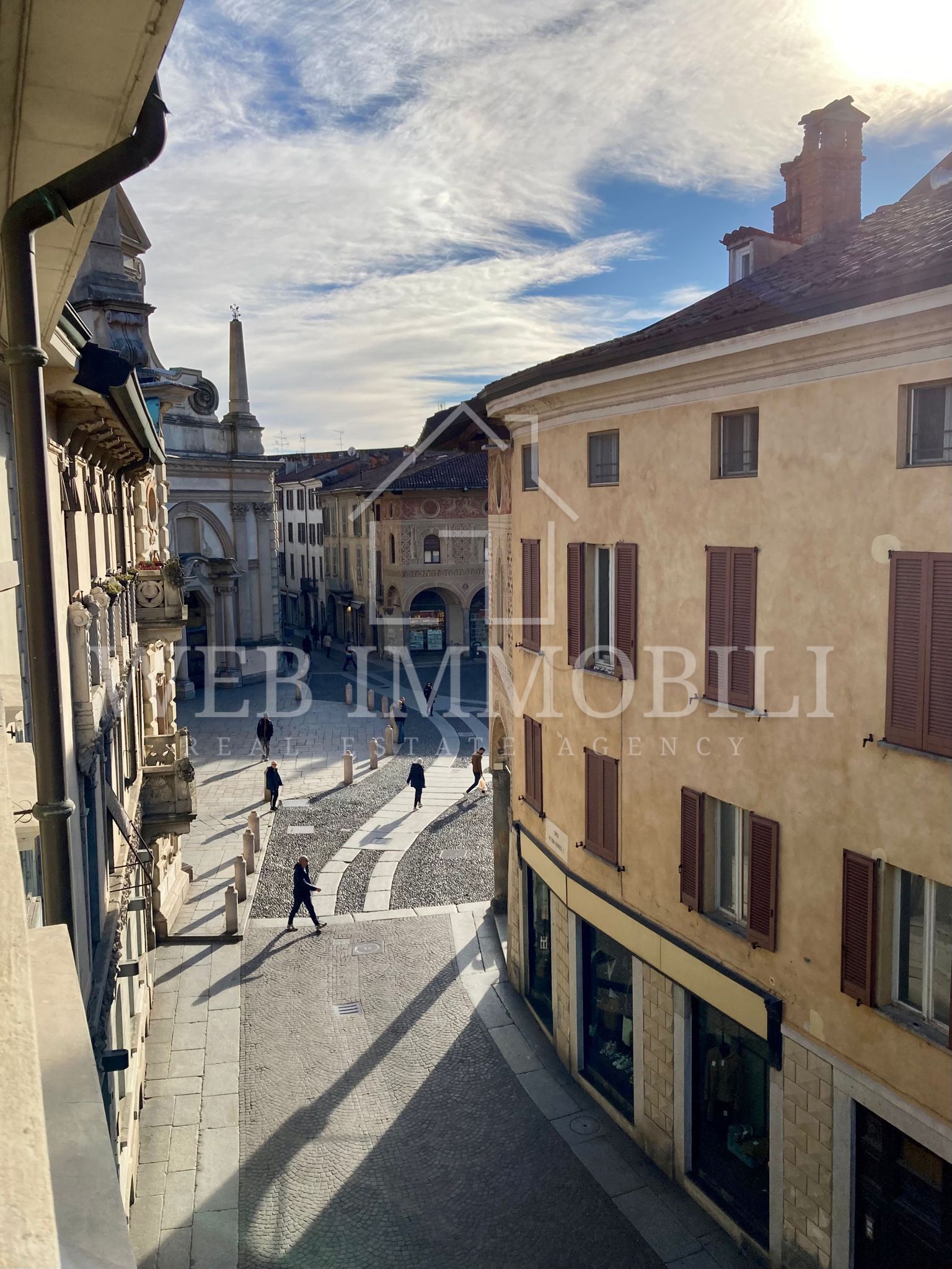 Veduta di una strada acciottolata con edifici, persone, un arco e una colonna in lontananza.