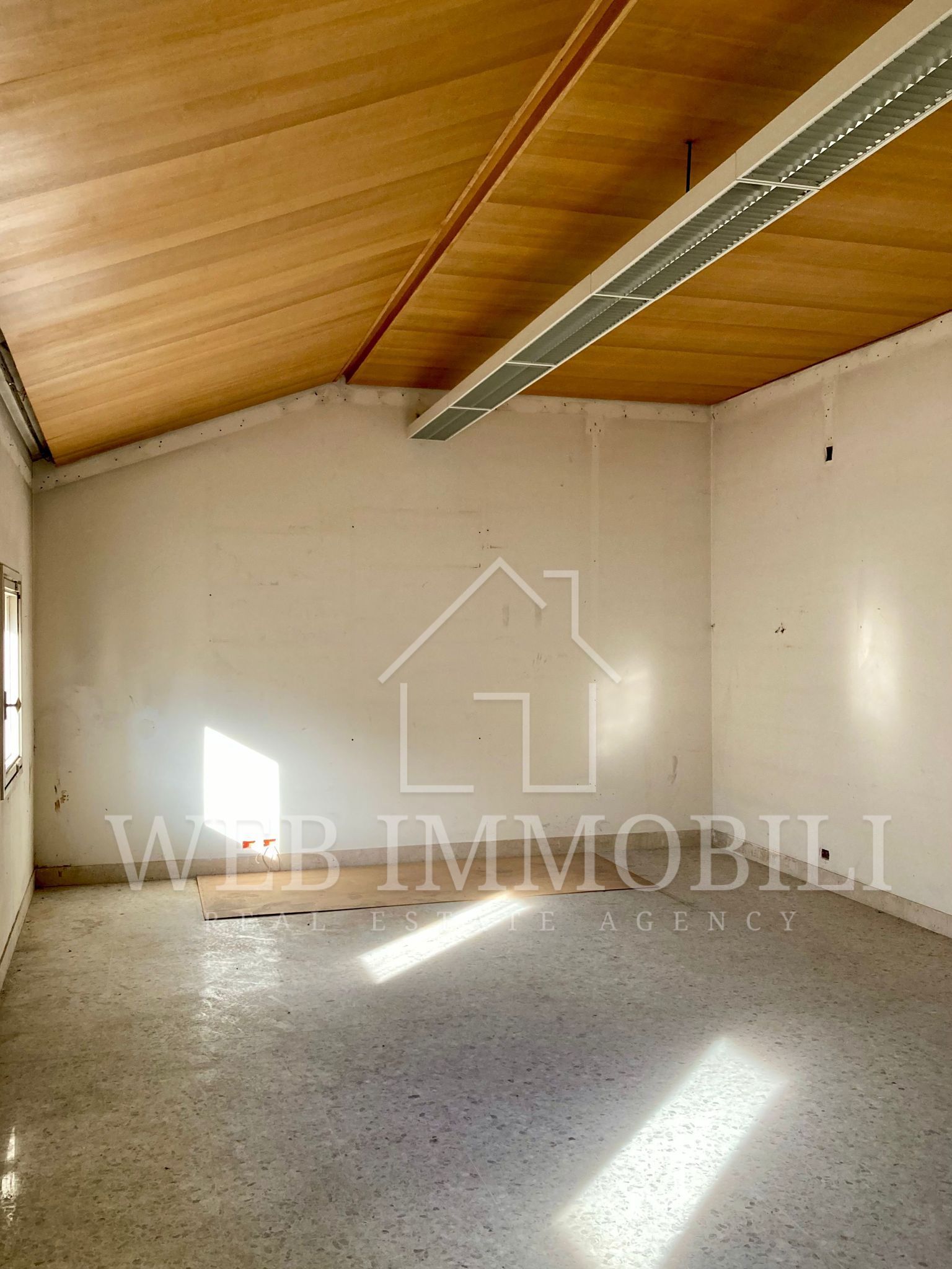 Stanza interna vuota con pareti chiare, soffitto in legno e pavimento punteggiato. La luce del sole filtra all'interno.