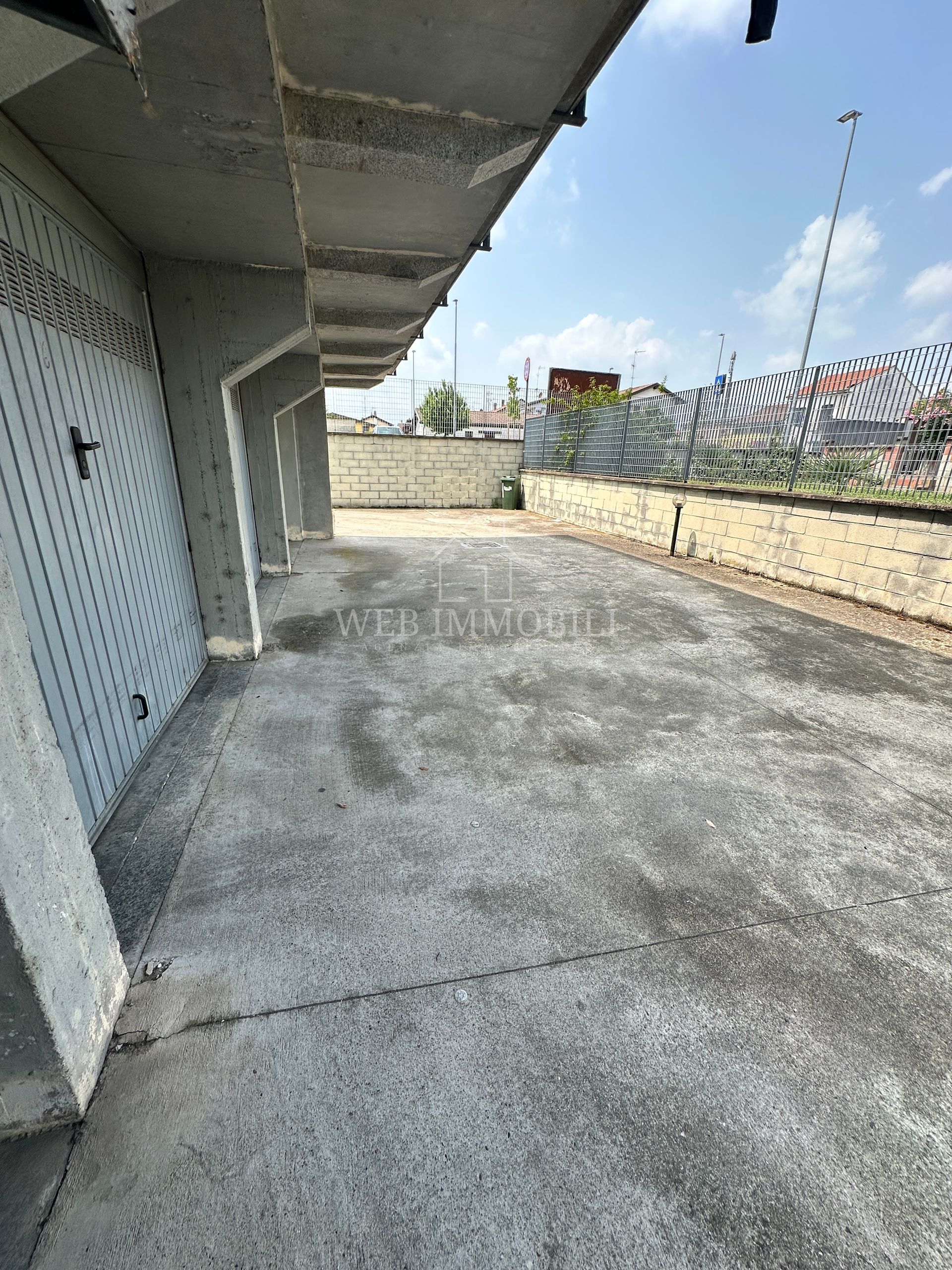 Fila di garage in cemento con porte grigie, sotto un edificio, giornata di sole.