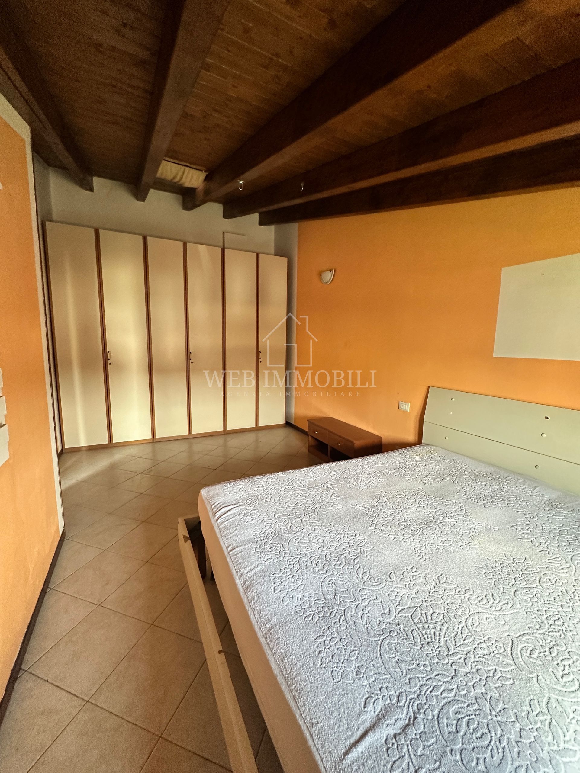 Camera da letto con parete arancione, armadio bianco, travi in ​​legno al soffitto e letto con piumone blu.