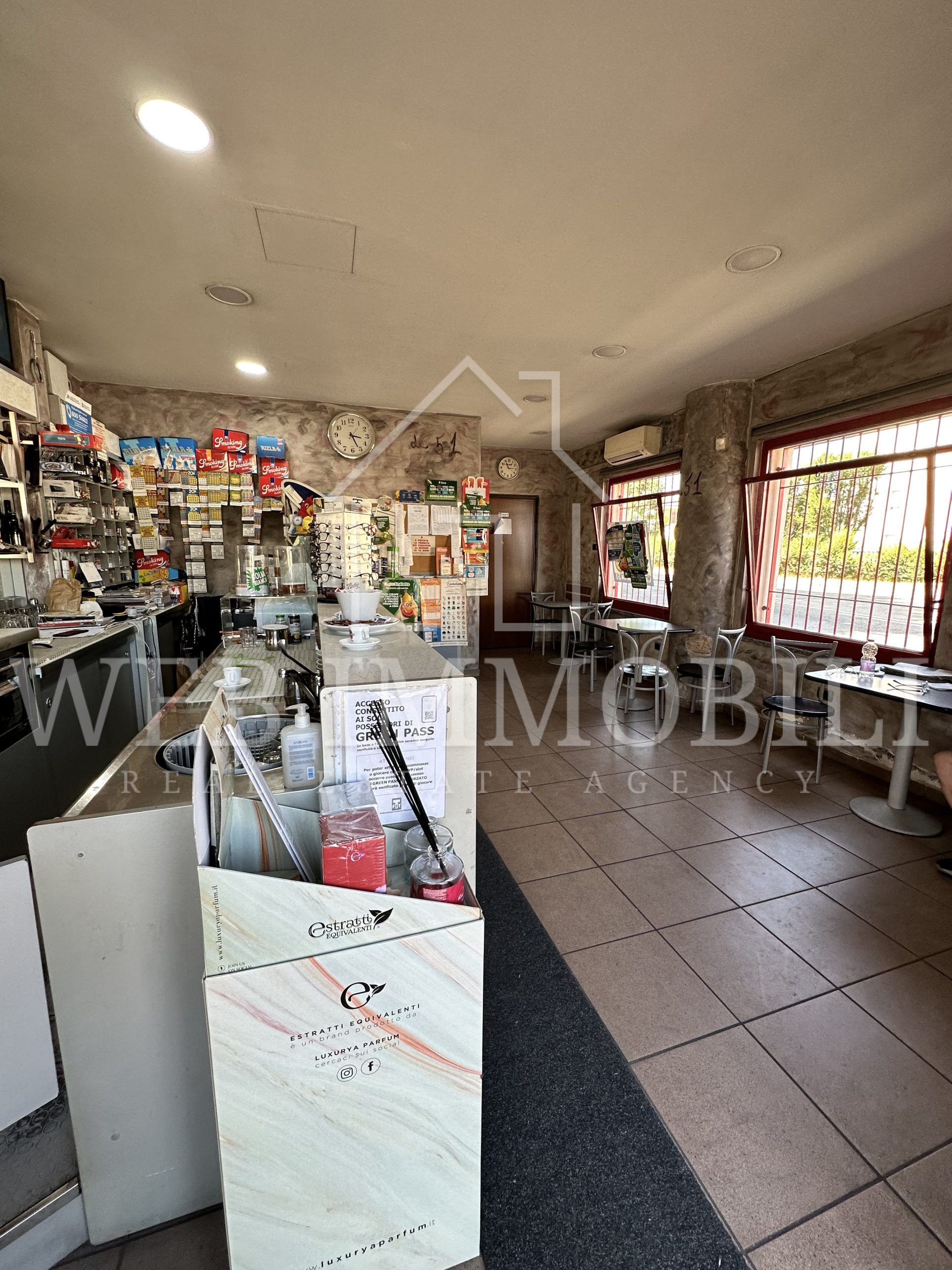 Interno di un bar con bancone, tavoli e porta. Pareti in pietra, pavimento piastrellato e merce a vista.