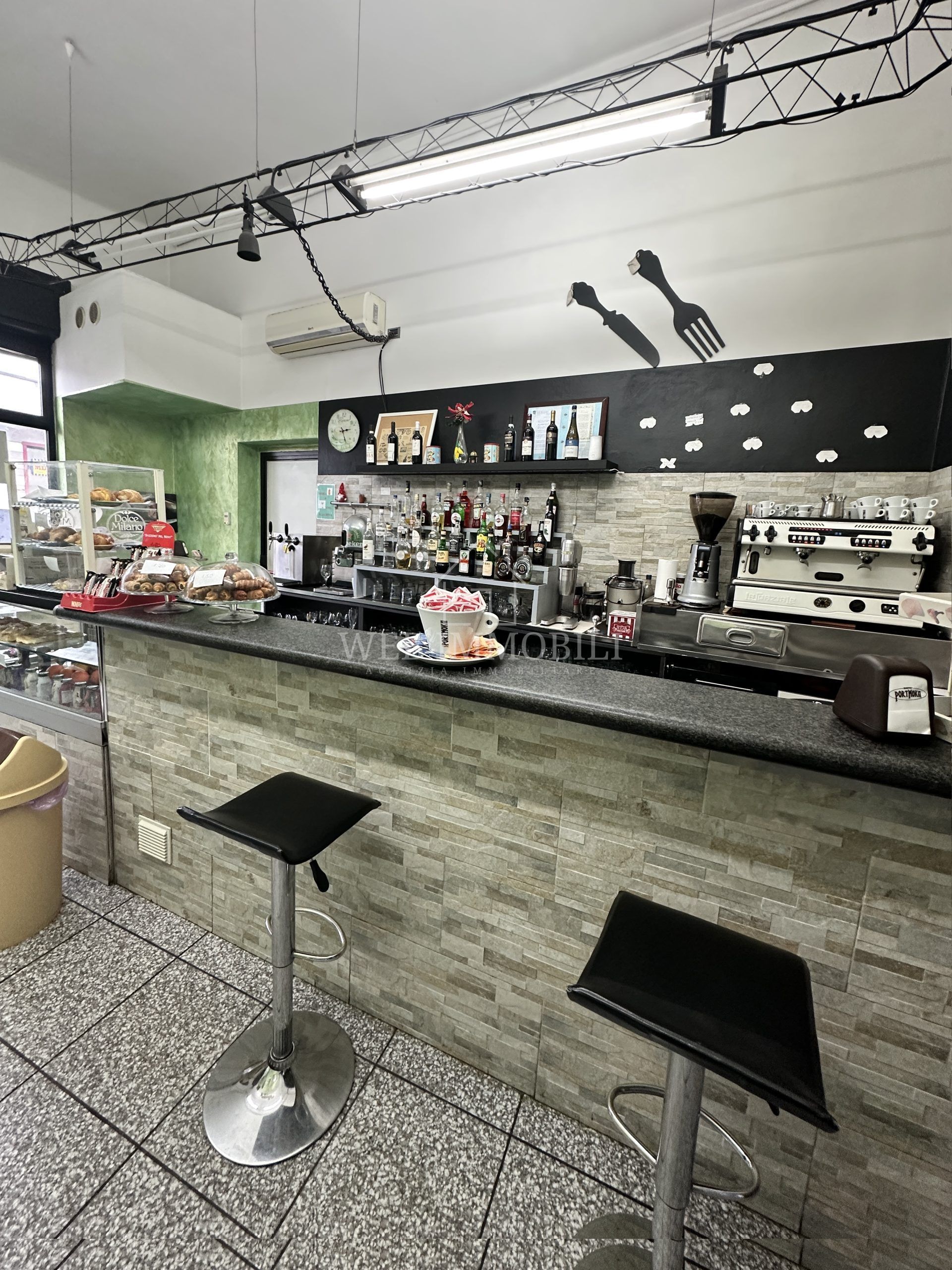 Interno di un bar con bancone, sgabelli, bevande e pasticcini.