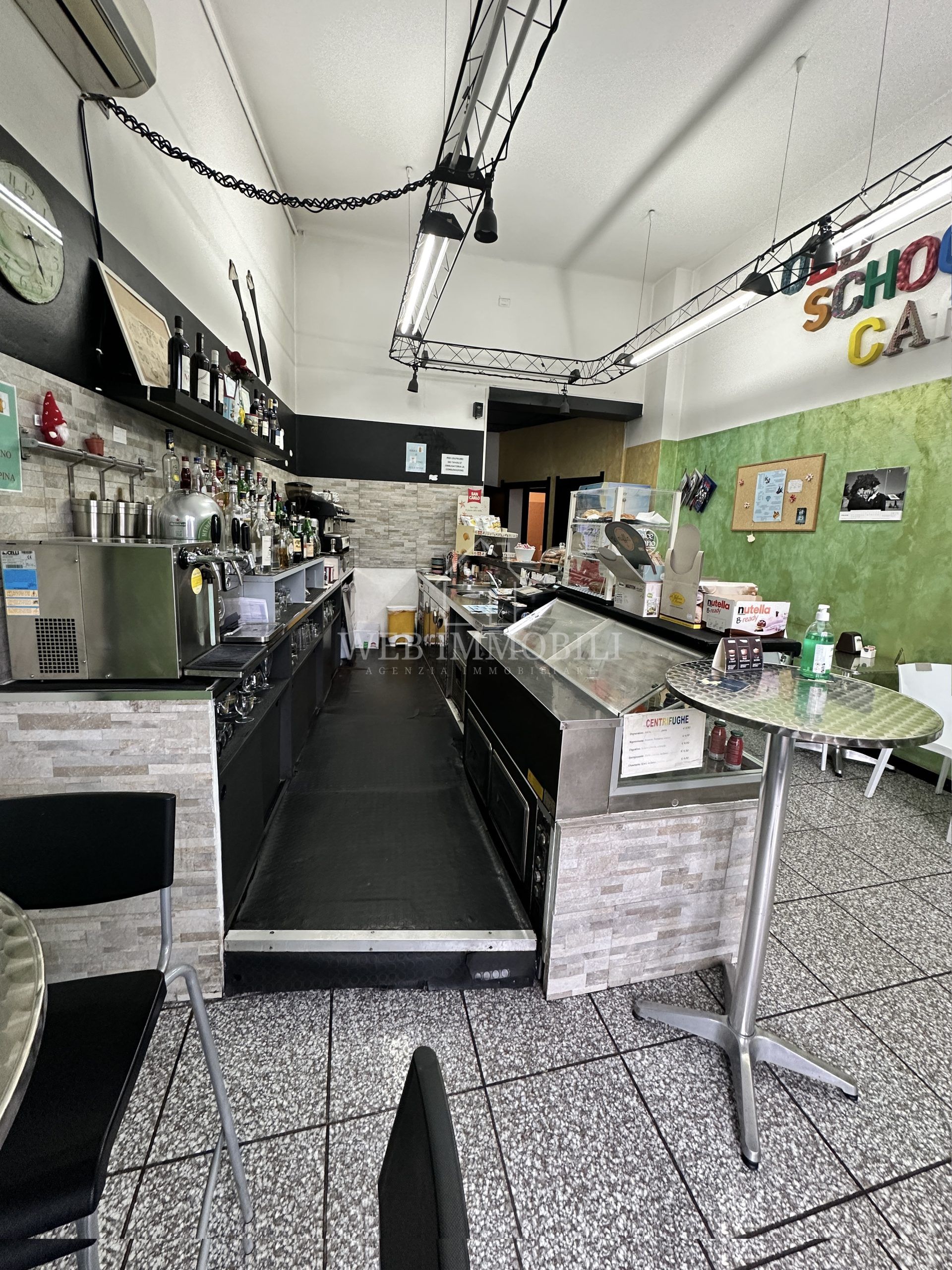 Interno di un bar/caffetteria con bancone, tavoli e merce. Pavimento piastrellato in bianco e nero, illuminazione intensa.