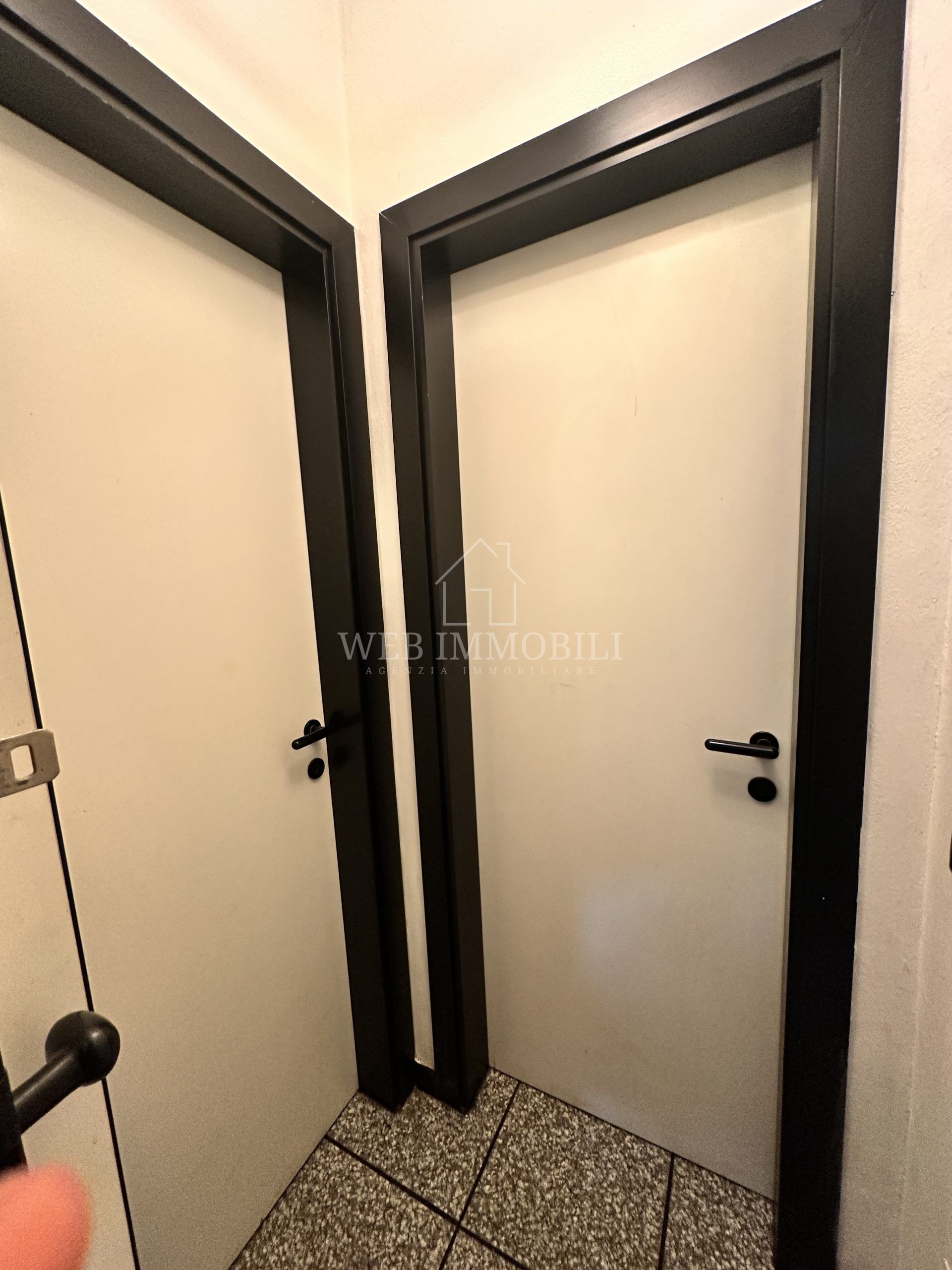 Due porte bianche con cornici e maniglie nere, inserite in un corridoio piastrellato.
