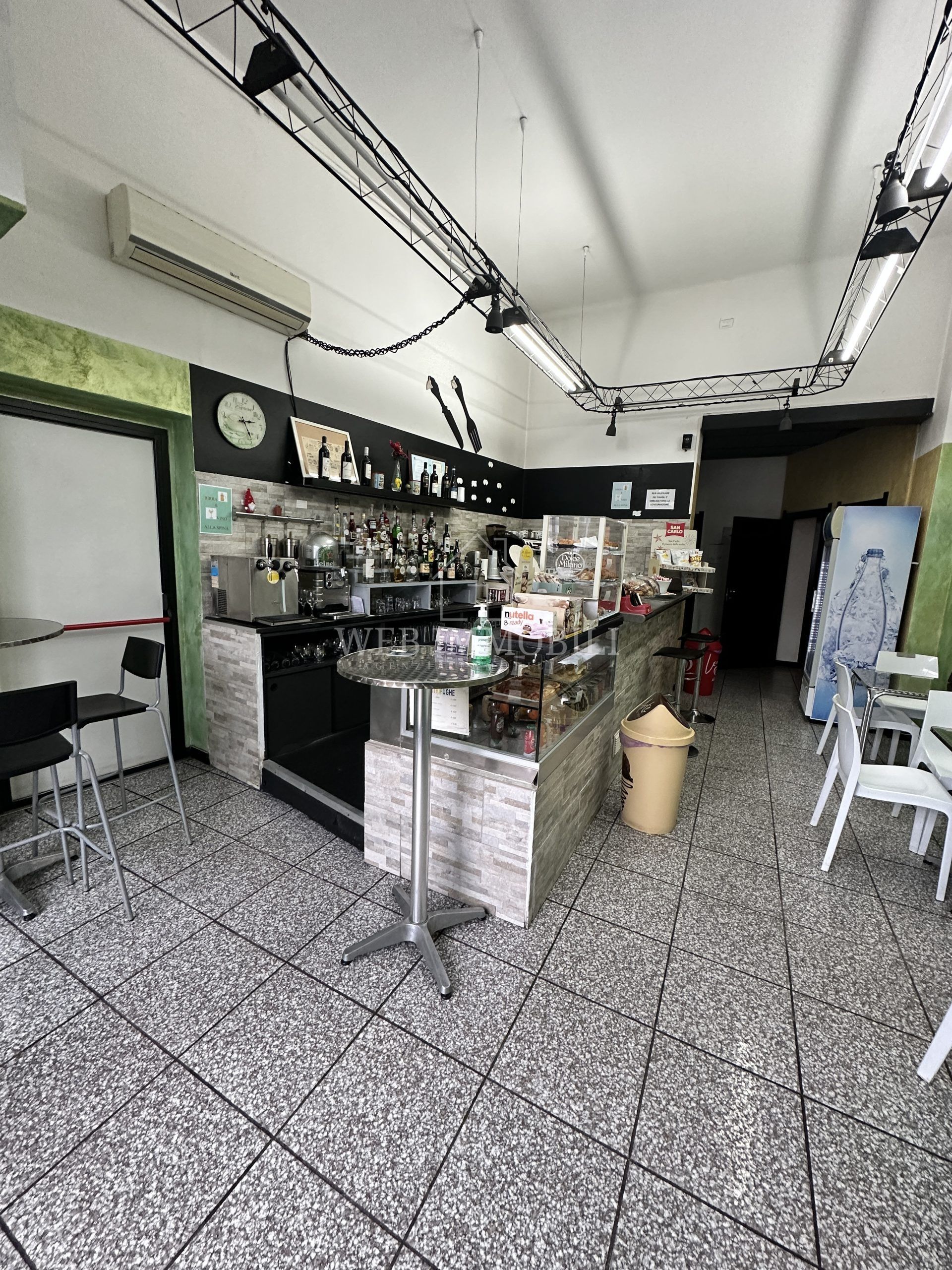 Interno di un bar con bancone, tavoli, sedie e vari prodotti alimentari esposti.