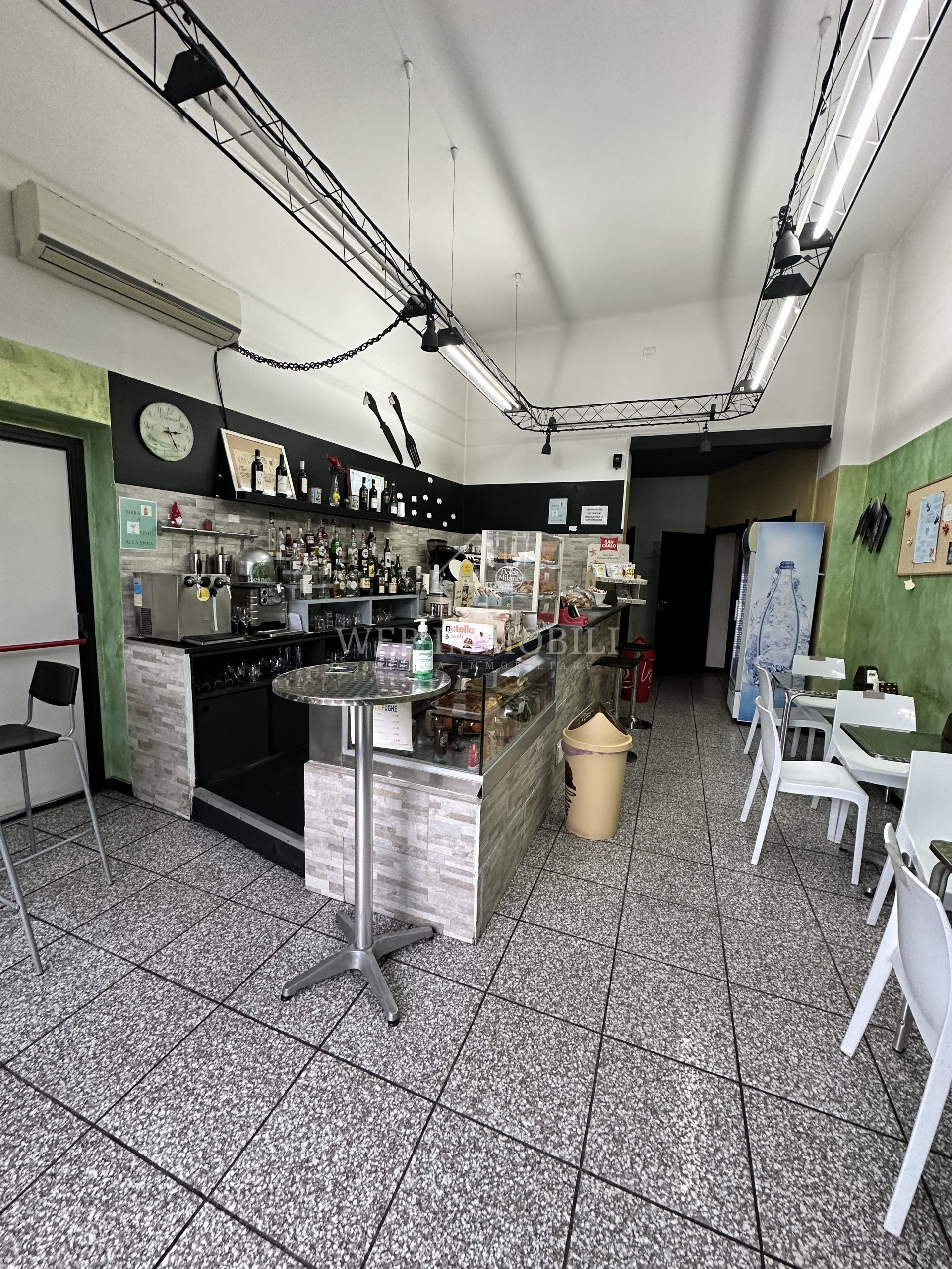 Interno di un piccolo bar con bancone, tavoli e sedie. Le pareti sono verdi e il pavimento è piastrellato.