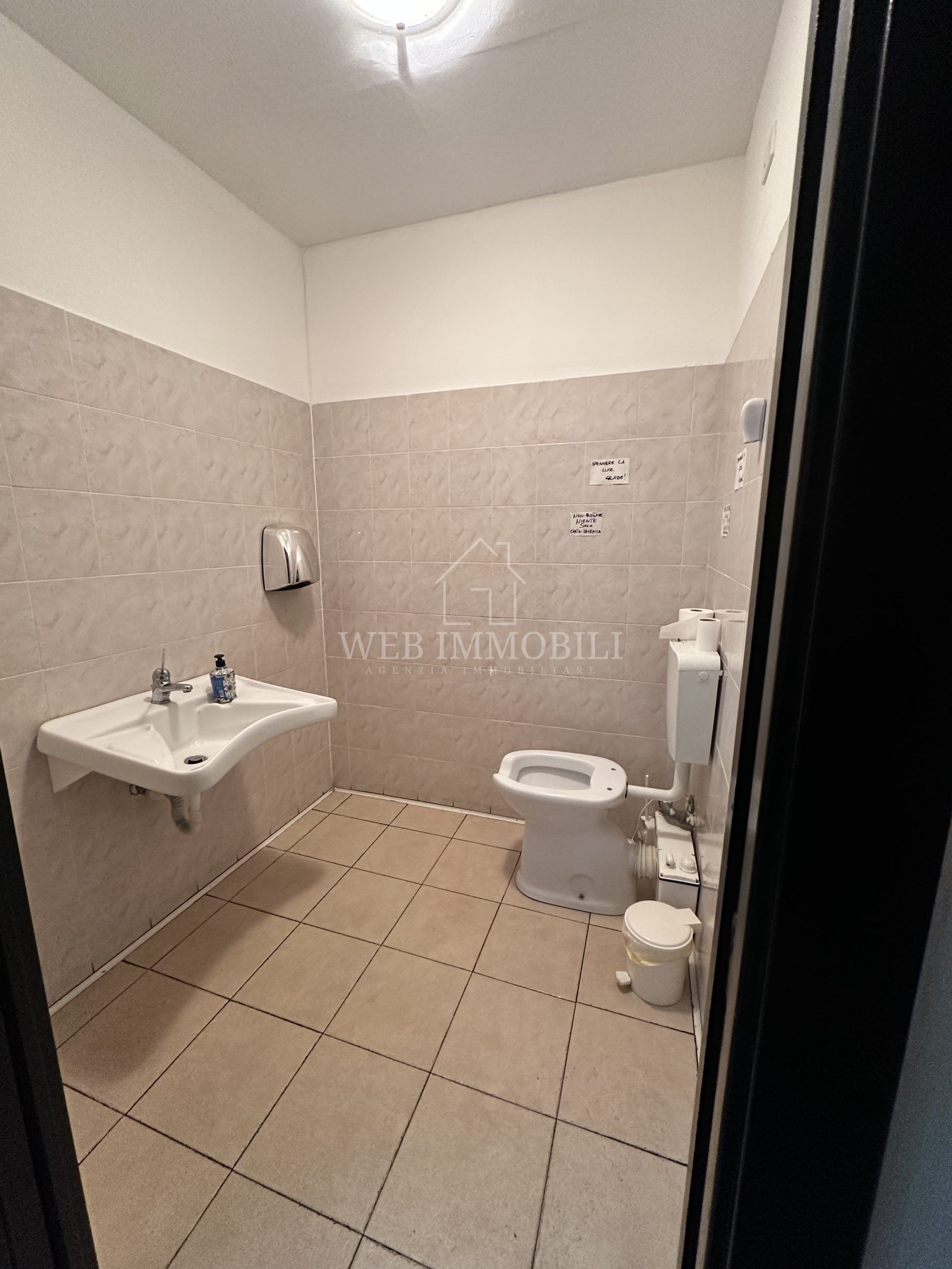 Piccolo bagno pubblico con lavandino, WC e asciugamani. Pareti beige e pavimento piastrellato.