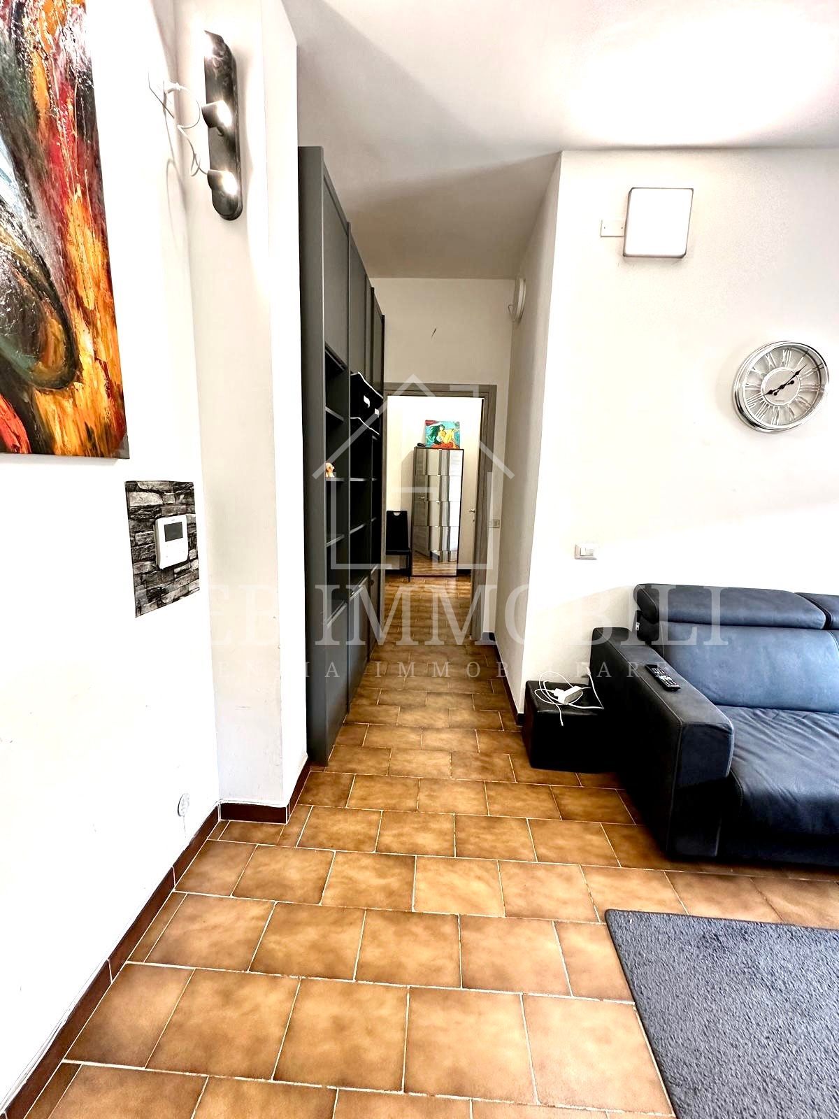 Corridoio stretto con pavimento piastrellato, mobili grigio scuro, pareti bianche, divano e opere d'arte.