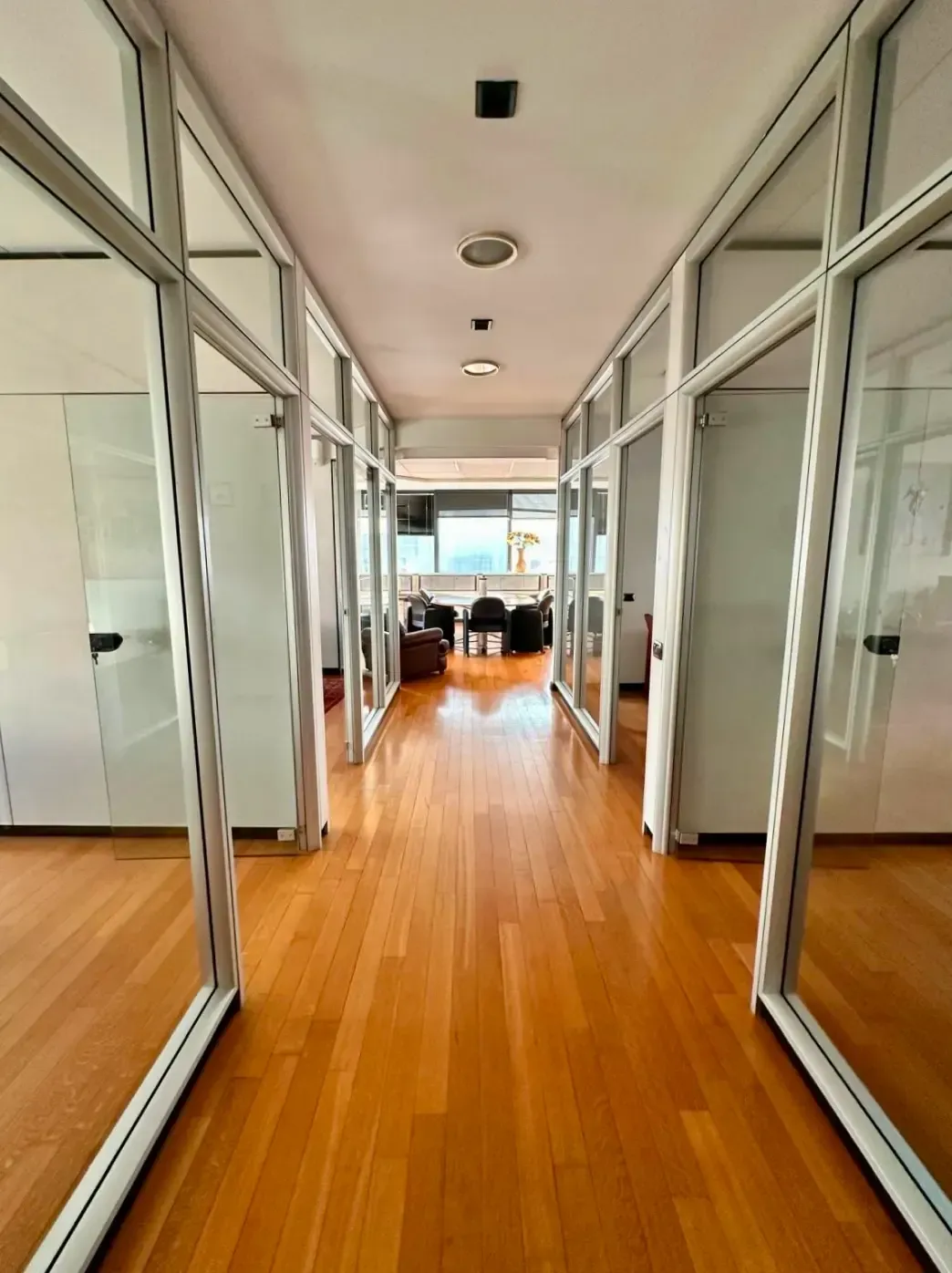 Corridoio con uffici con pareti in vetro, pavimento in legno e luci zenitali; vista verso la fine del corridoio.