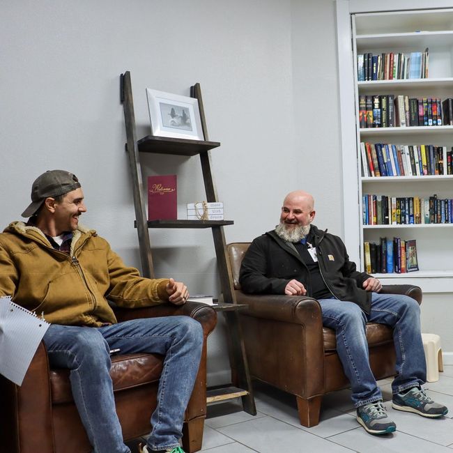 Two men are sitting in chairs in front of a bookshelf