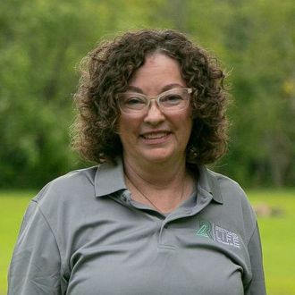 A woman with curly hair and glasses is wearing a gray shirt and smiling.