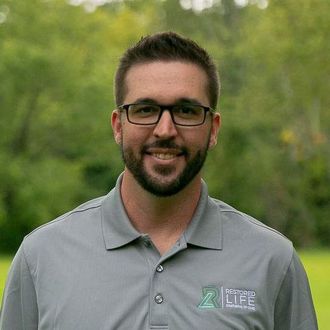 A man with glasses and a beard is wearing a grey shirt and smiling for the camera.