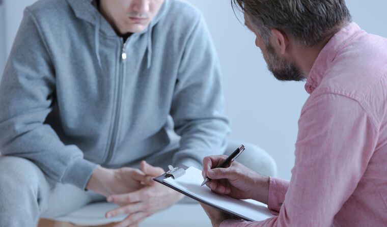 A man is sitting on the floor talking to another man while holding a clipboard.