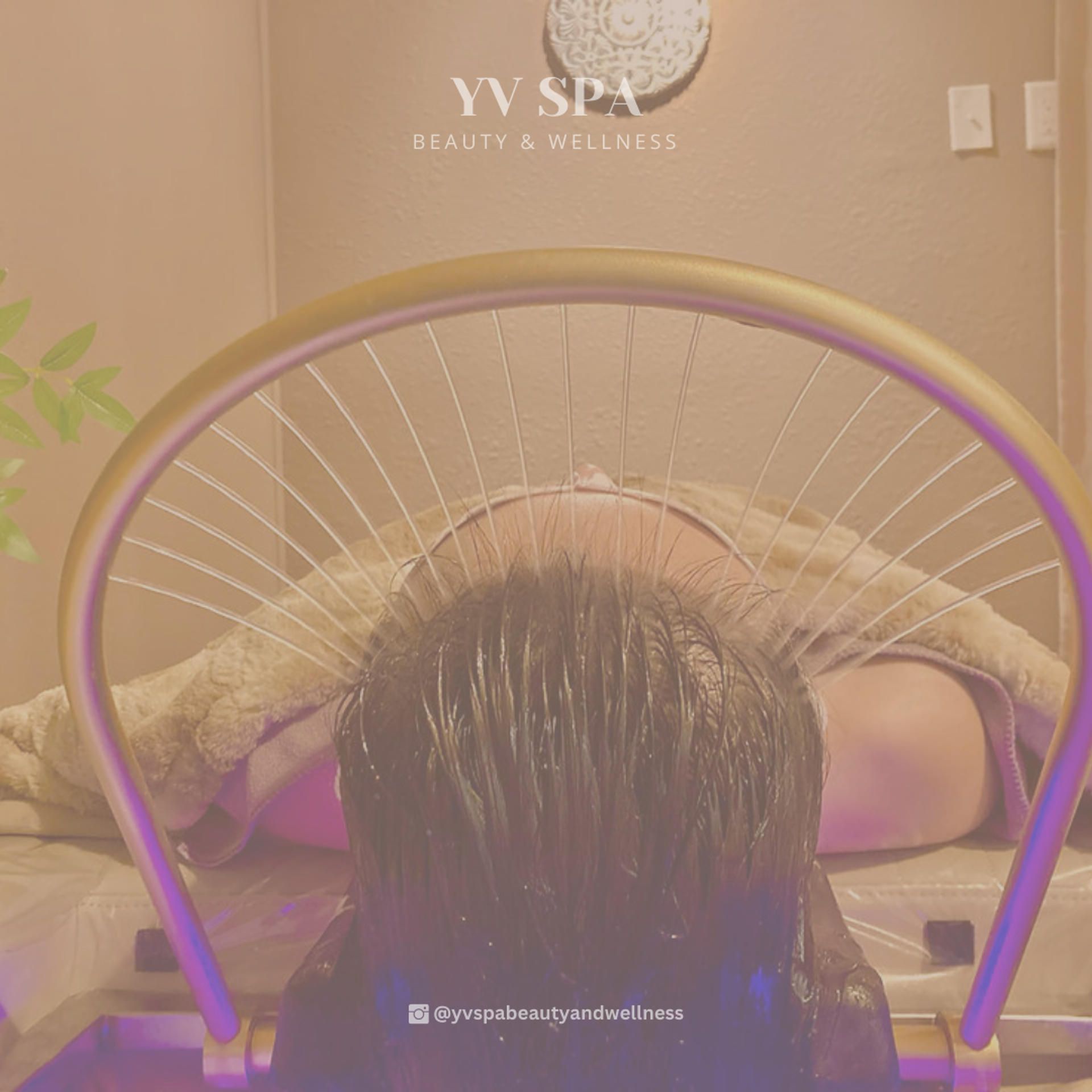 Person receiving hair treatment at a spa, beneath a curved metallic device, in a beige room.
